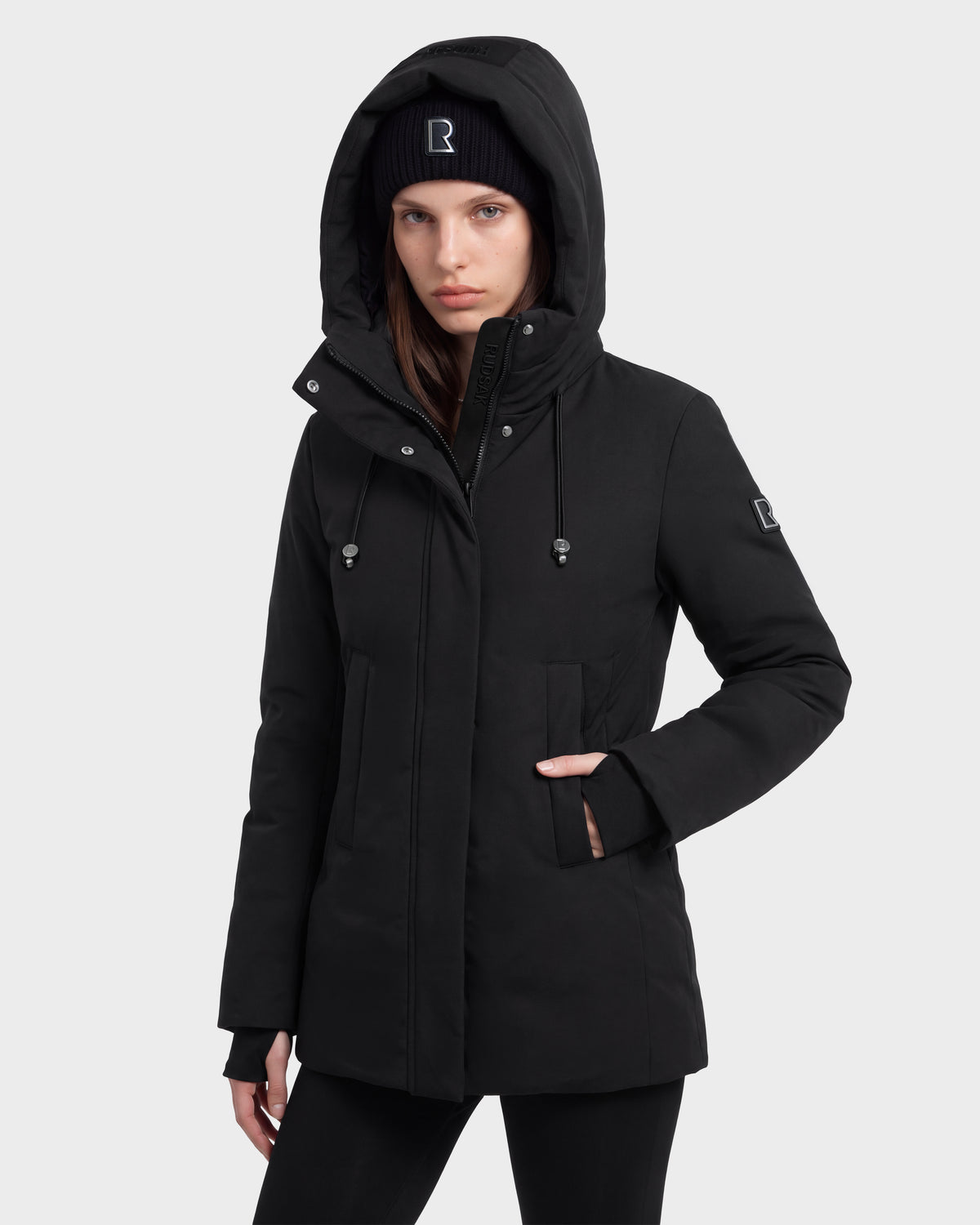 A person stands facing forward with hands in the pockets of a Rudsak OPAL - 8126618 Black hooded winter jacket and a matching black beanie, both featuring a small white logo, against a plain light background.