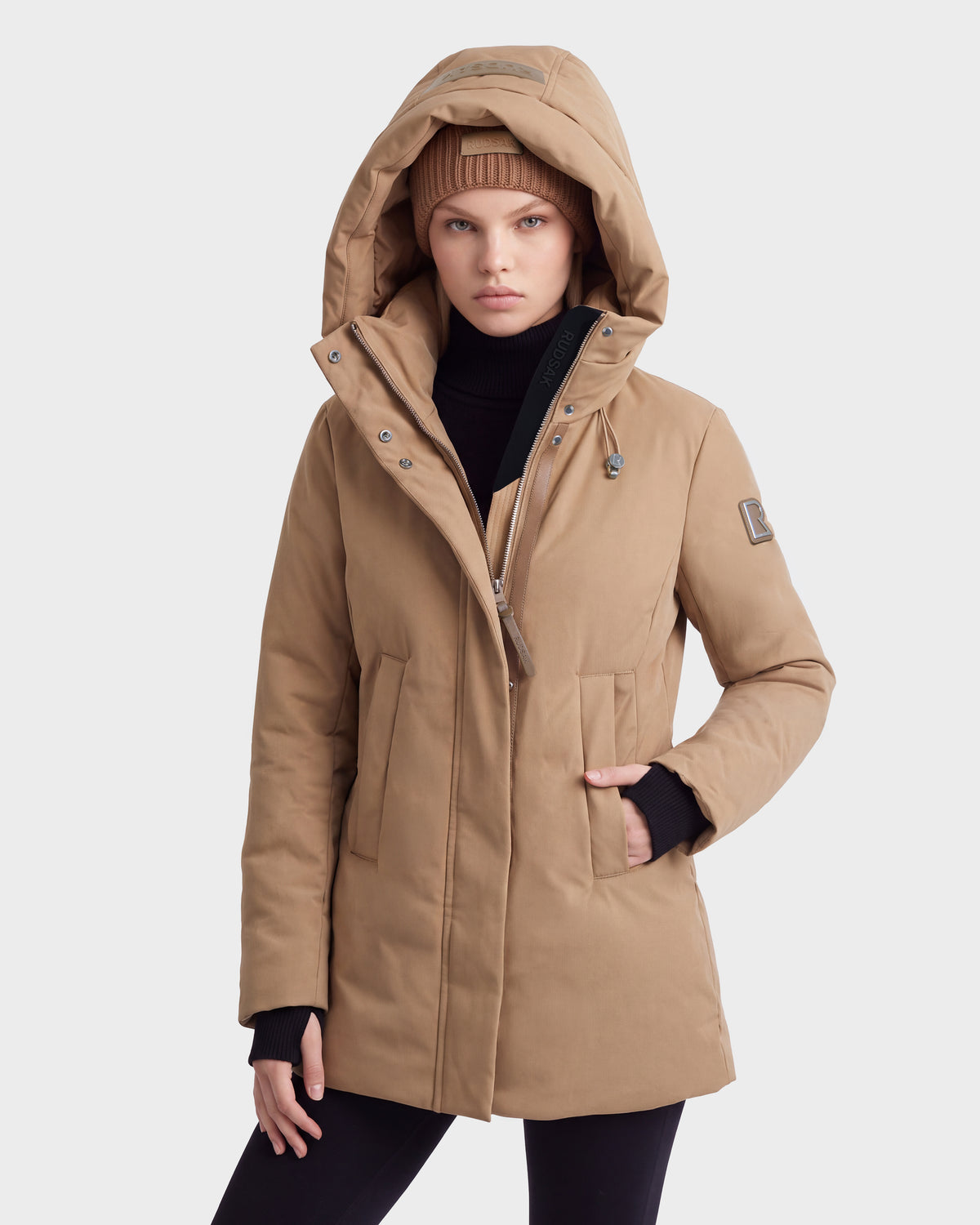 Someone wearing the OPAL - 8126618 Camel by Rudsak and a matching knit hat poses with one hand in their pocket against a plain light background.
