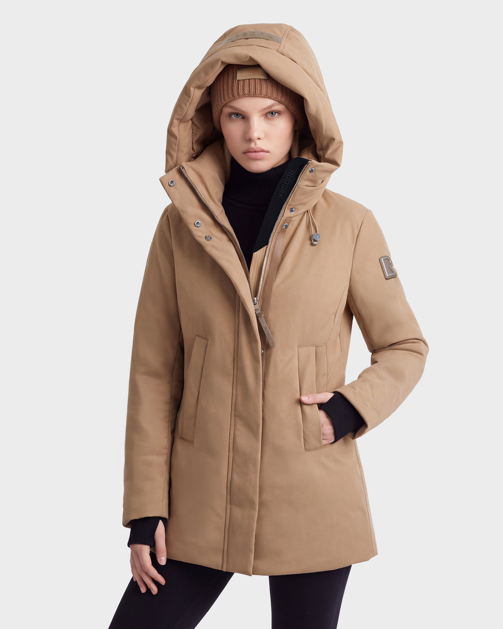 Someone models the OPAL - 8126618 Camel by Rudsak, a tan hooded parka with the hood up and a matching beanie, one hand in their pocket against a plain light background.