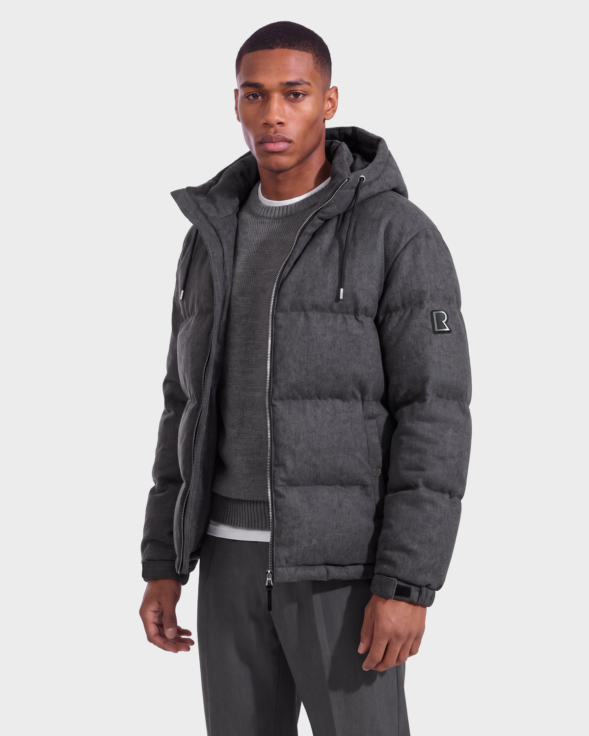 A man wearing the Rudsak STERLING W - 6126760 Heather Charcoal hooded puffer jacket over a gray sweater and gray pants stands facing forward against a plain light background.