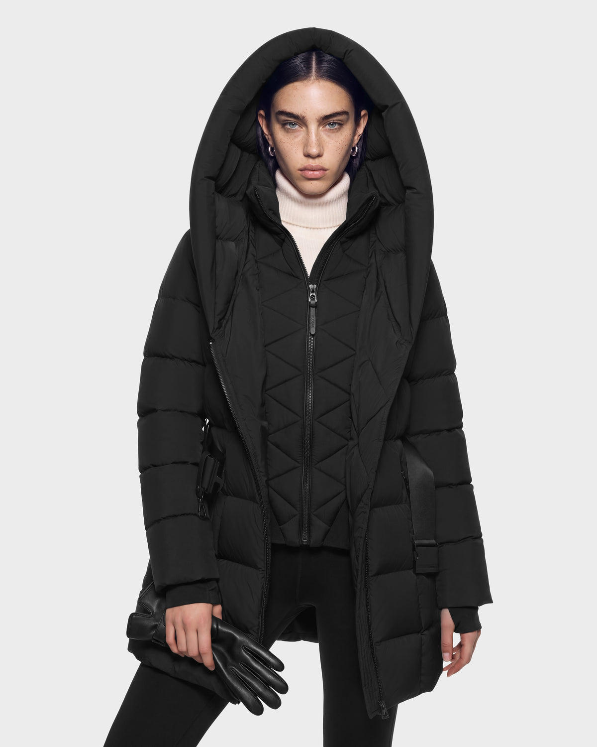 A person in a black hooded, fitted quilted silhouette Rudsak TAYLOR M - 8125965 Black puffer coat over a white turtleneck sweater, holding a pair of black gloves.