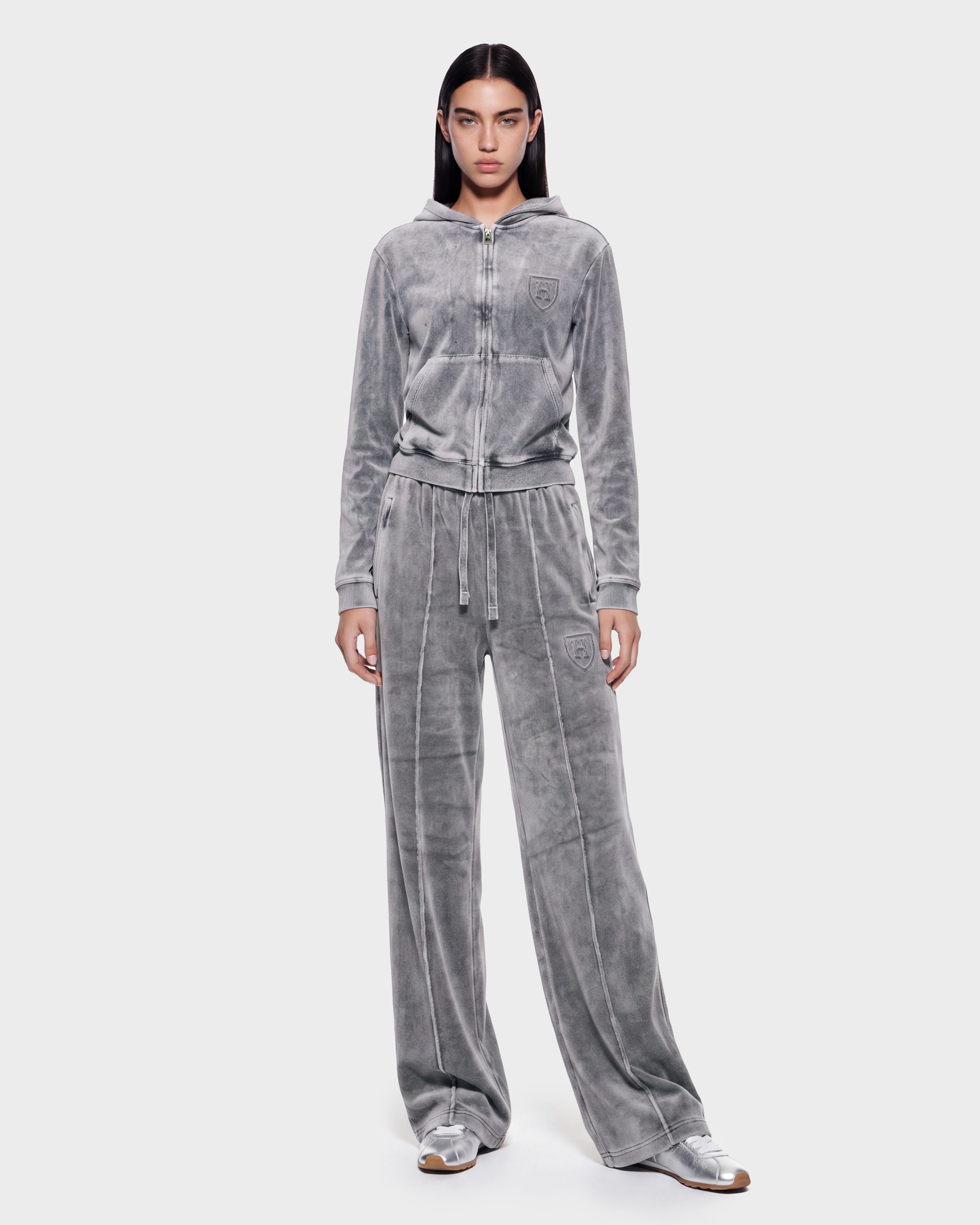 A person stands facing forward against a plain light background, wearing the Rudsak TYRA HV - H526904 Titanium gray velour tracksuit with a zip-up hoodie and wide-leg pants, paired with silver sneakers.