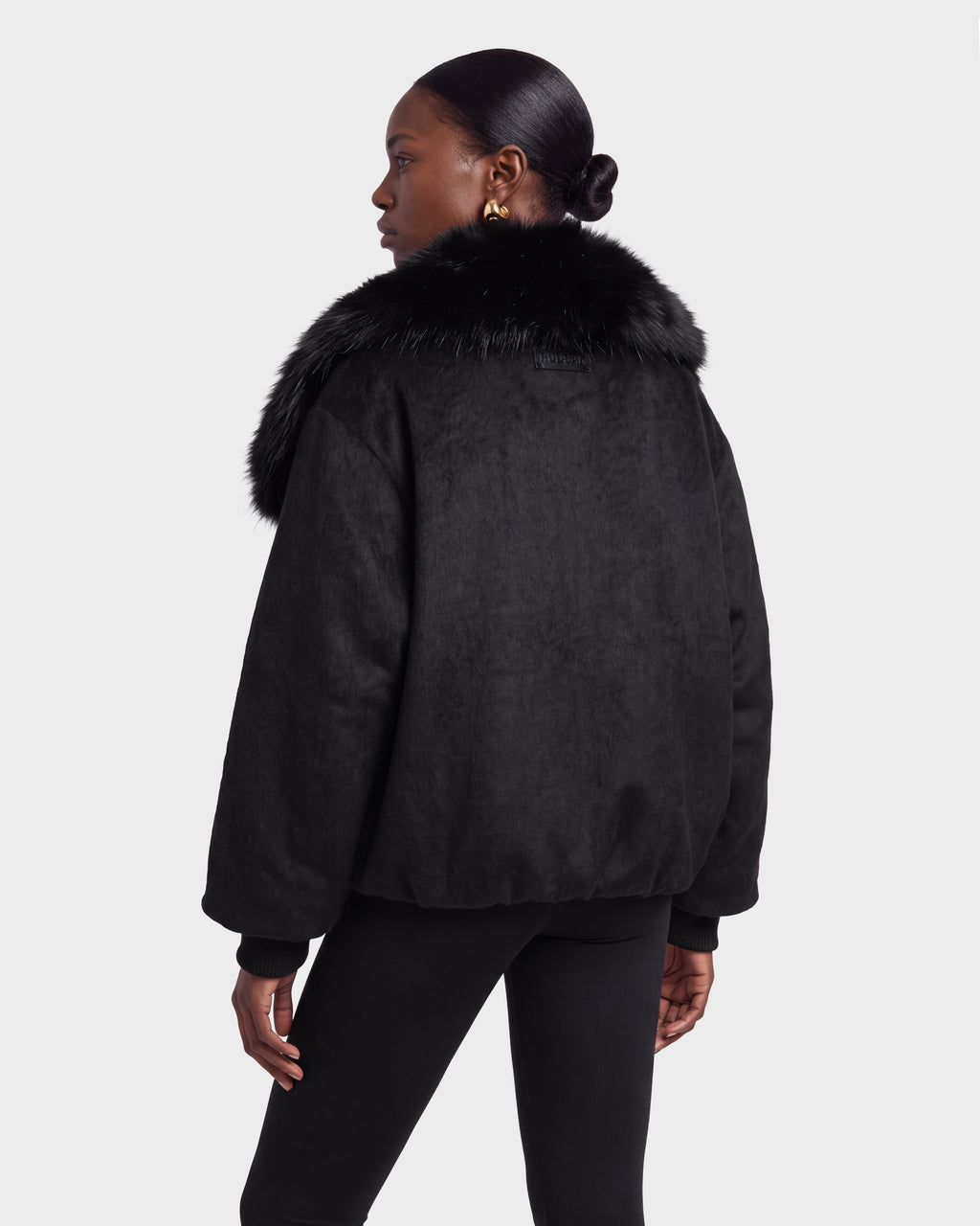 A woman stands facing away, looking to the side against a plain background, wearing the Rudsak ZARIA - 8126999 Black faux fur-collared jacket and black pants.