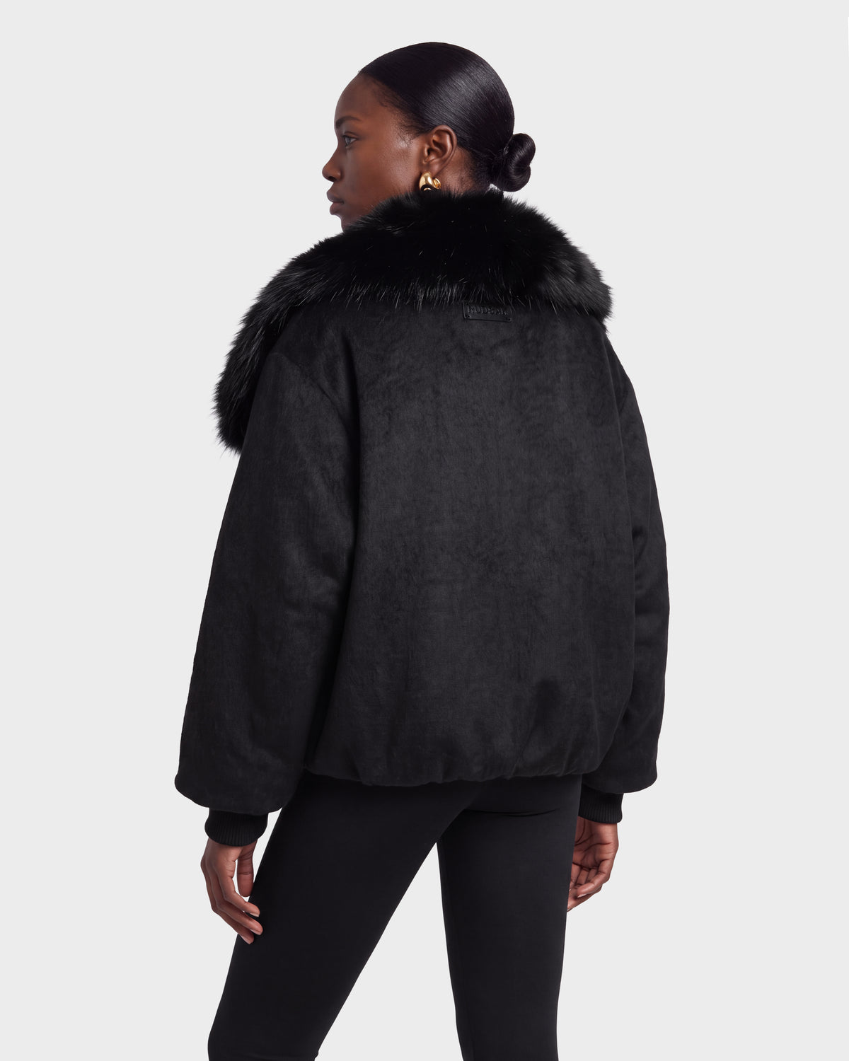 A woman stands facing away, looking to the side against a plain background, wearing the Rudsak ZARIA - 8126999 Black faux fur-collared jacket and black pants.