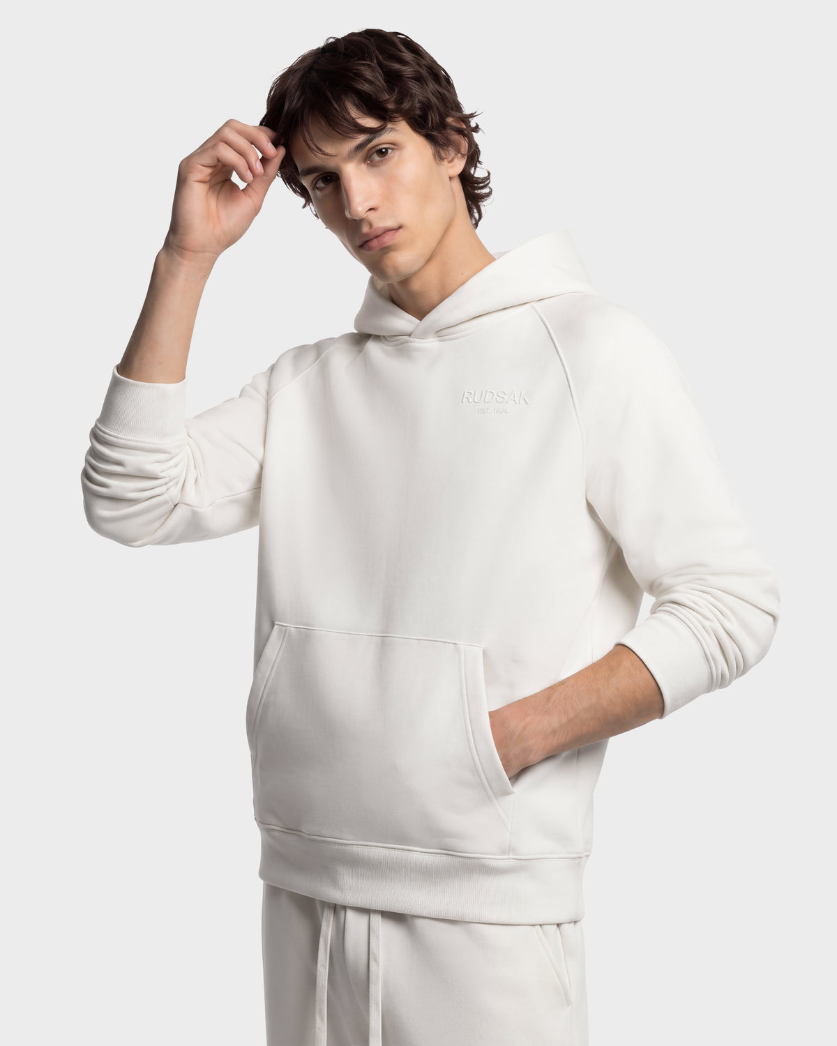 A person wearing a cream-colored RUDSAK BRIAR - 9525001 Vanilla M unisex hoodie with a front kangaroo pocket and a small logo on the chest.