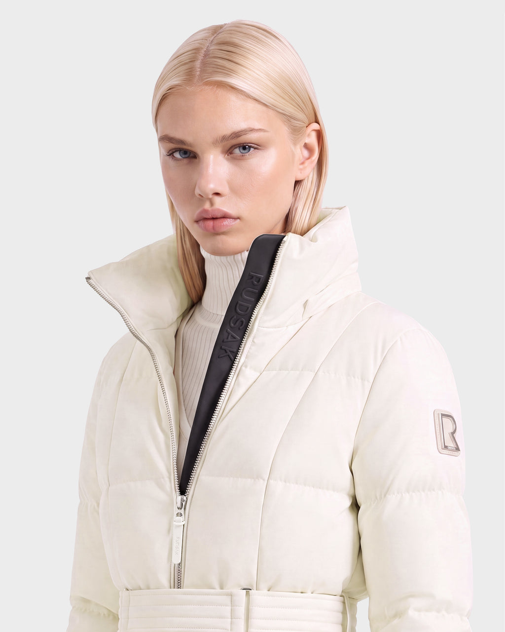 A woman wears a knee-length, vanilla-colored Rudsak AUDREY L - 8124585 Vanilla puffer jacket with a high collar and a matching belt.