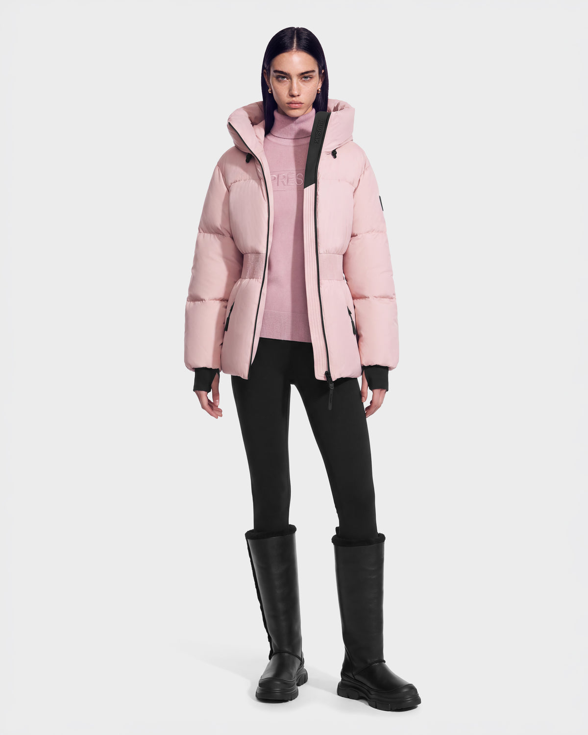 A person wearing a Rudsak DIAMOND - 8126772 Vintage Rose pale pink quilted puffer jacket with a hood, a matching turtleneck sweater, black leggings, and black knee-high boots.