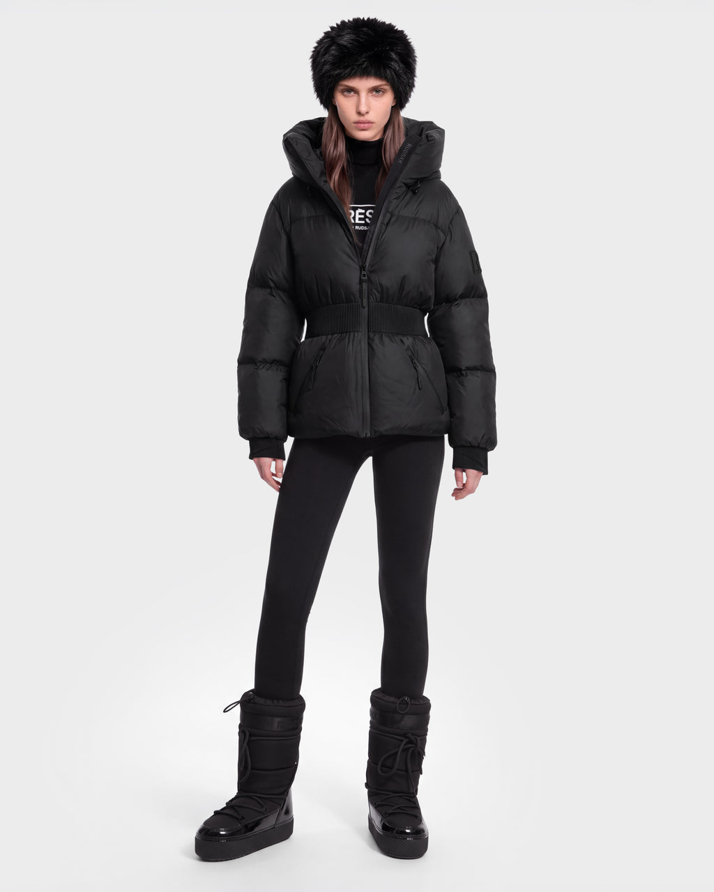 A person wears the Rudsak DIAMOND - 8126772 Black puffer jacket, black leggings, winter boots, and a black fur hat while standing against a plain white background.