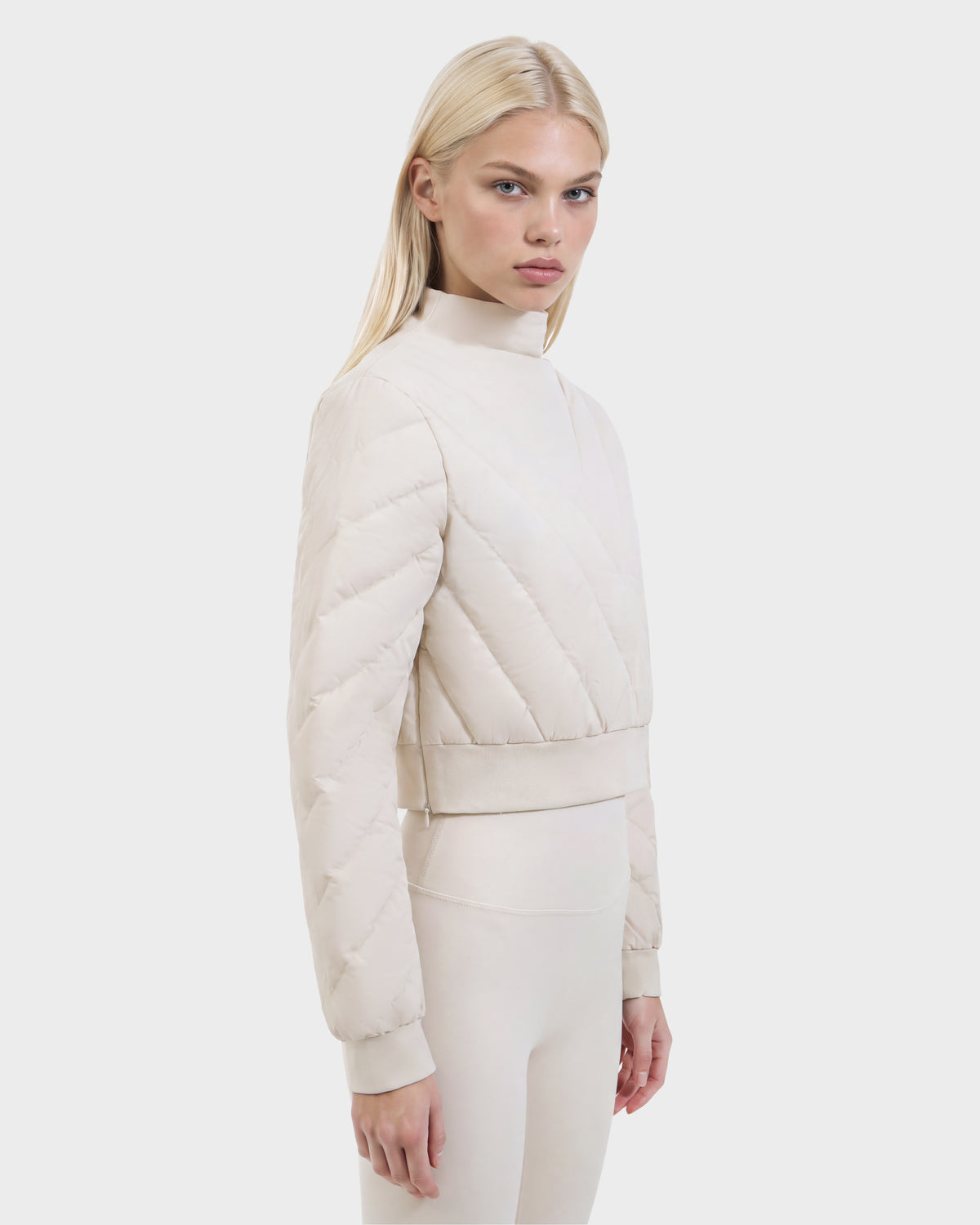 A blonde woman wears the Rudsak KATYA - 8526531 Vanilla, a cropped, vanilla-colored quilted jacket with a mock neck, styled as an après-ski layer.