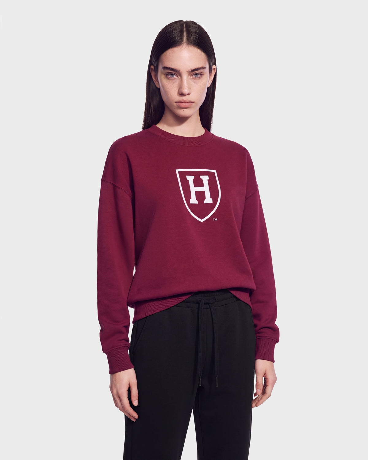 A person with straight brown hair wears the Rudsak HARVARD ONE CRIMSON SWEATSHIRT 9524507—a maroon unisex sweater adorned with a white shield and H logo—paired with black pants and standing against a plain gray background, capturing classic Ivy League style.