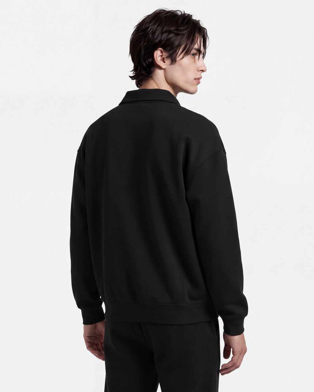 A person with medium-length dark hair models the Rudsak IDRIS - 6526500 Black jacket, featuring a polo collar, paired with black pants, standing against a plain white background and looking over their left shoulder.