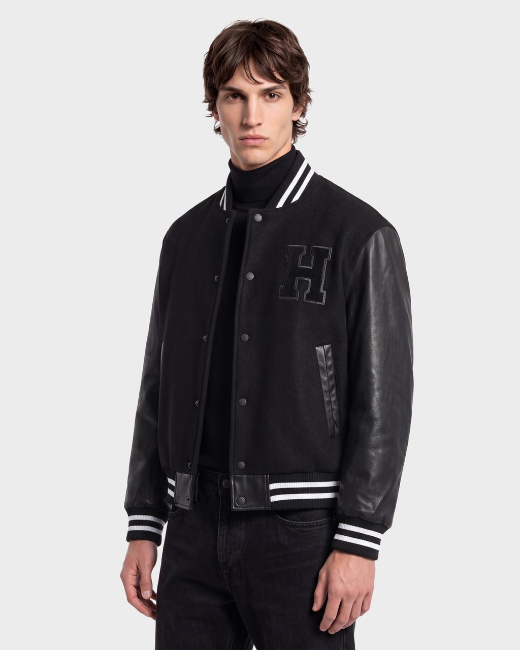 A young man in the Rudsak JOHN HV - H125504 Black jacket—a limited edition collegiate-inspired varsity jacket featuring leather sleeves and an H patch on the chest—stands against a plain light background, looking at the camera.
