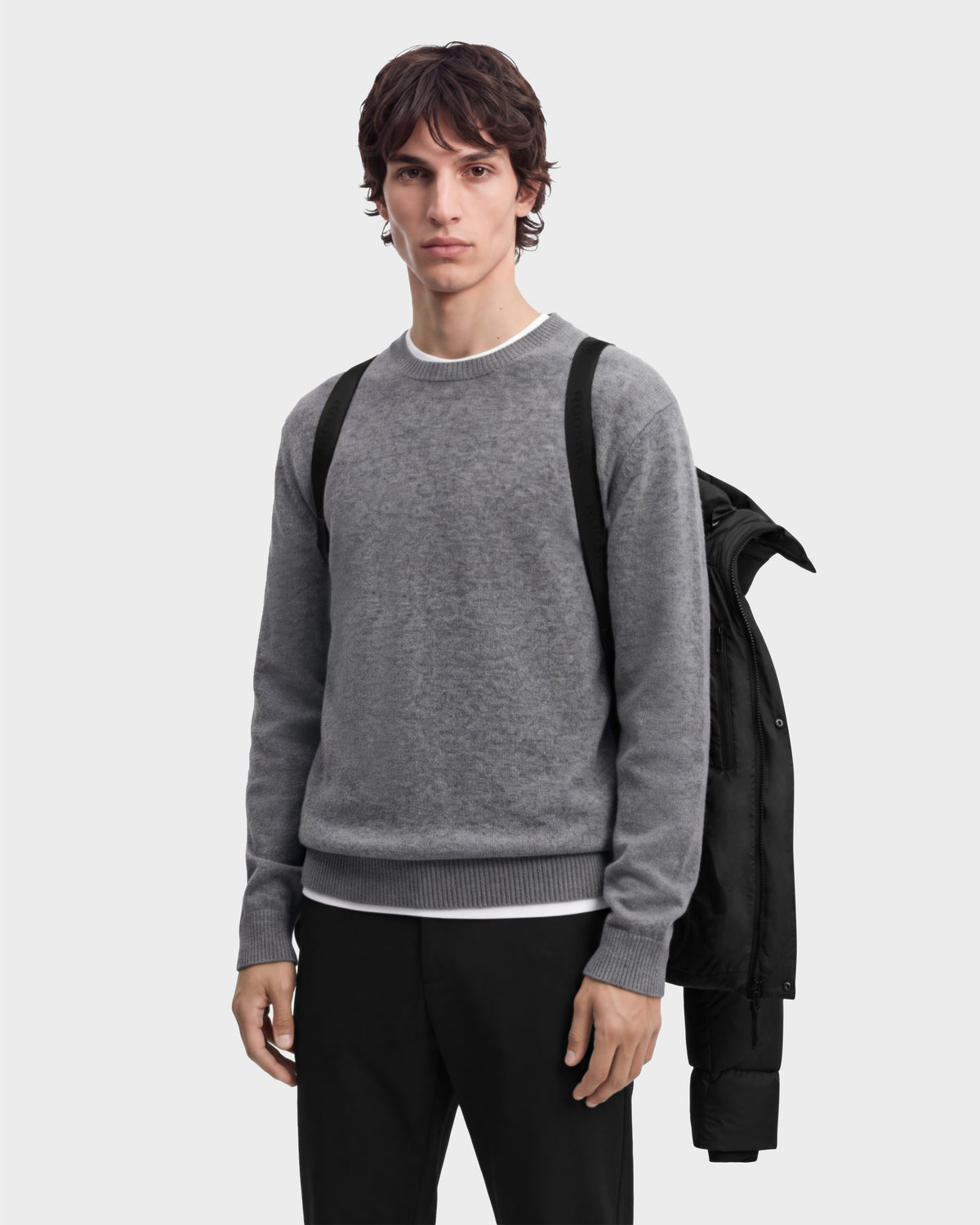 A person wearing a grey crewneck sweater over a white t-shirt and black trousers, with a black JASON - 6126803 puffer jacket draped over their shoulder.