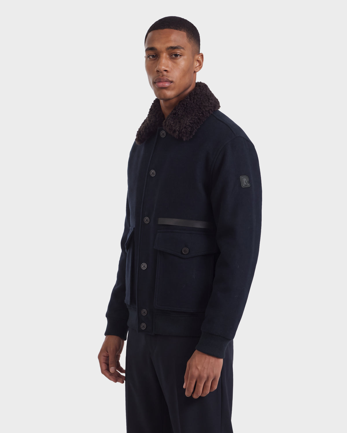 A man wearing a Rudsak JULIUS - 6126723 Navy wool-blend jacket with a dark faux-fur collar, buttoned front, and two large front flap pockets.