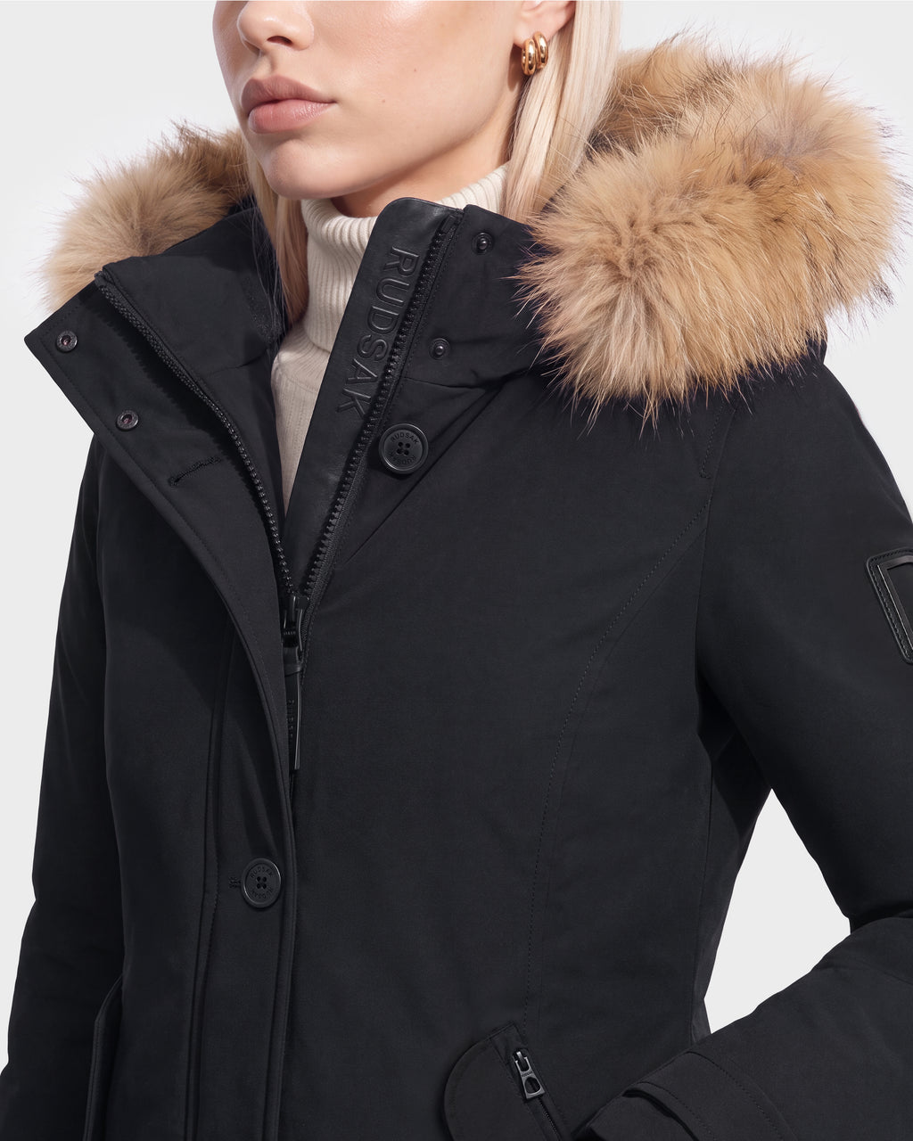 Woman in the Rudsak KEYSHIA - 8126517 Black winter coat with a tan faux fur-trimmed hood, white turtleneck, and gold hoops—showcasing a cold-weather essential against a plain background.