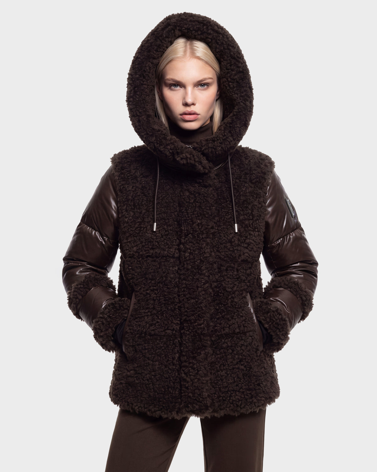 A person wearing the Rudsak MARISSA - 8126728 Espresso coat, a dark brown textured winter outerwear piece with leather sleeves and a hood, stands against a plain light gray background with hands in pockets, looking directly at the camera.