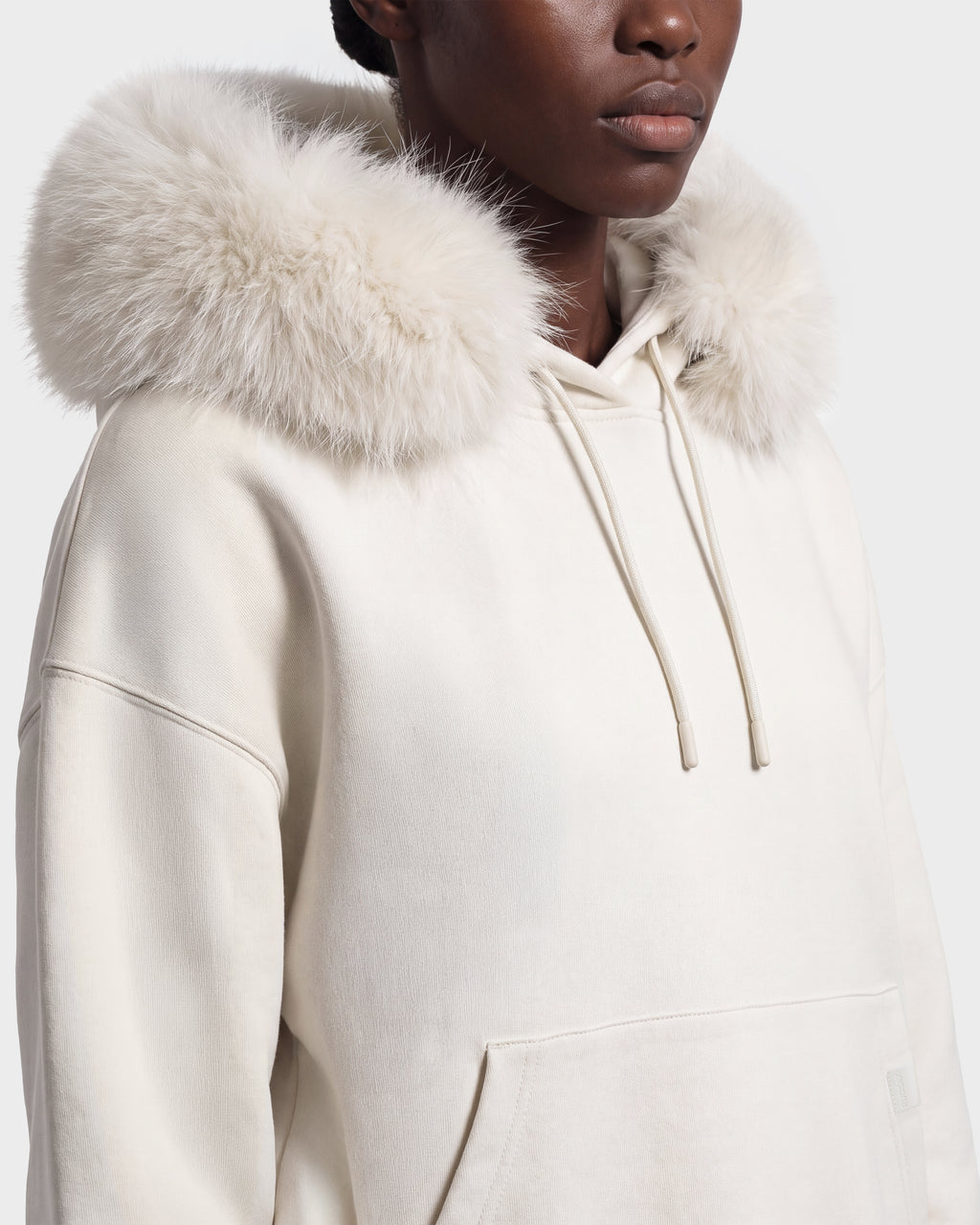 A person wears the Rudsak MILENA - 8526689 Vanilla, a cream-colored hoodie with a large, fluffy fur-trimmed hood, showing only their lower face and upper torso.