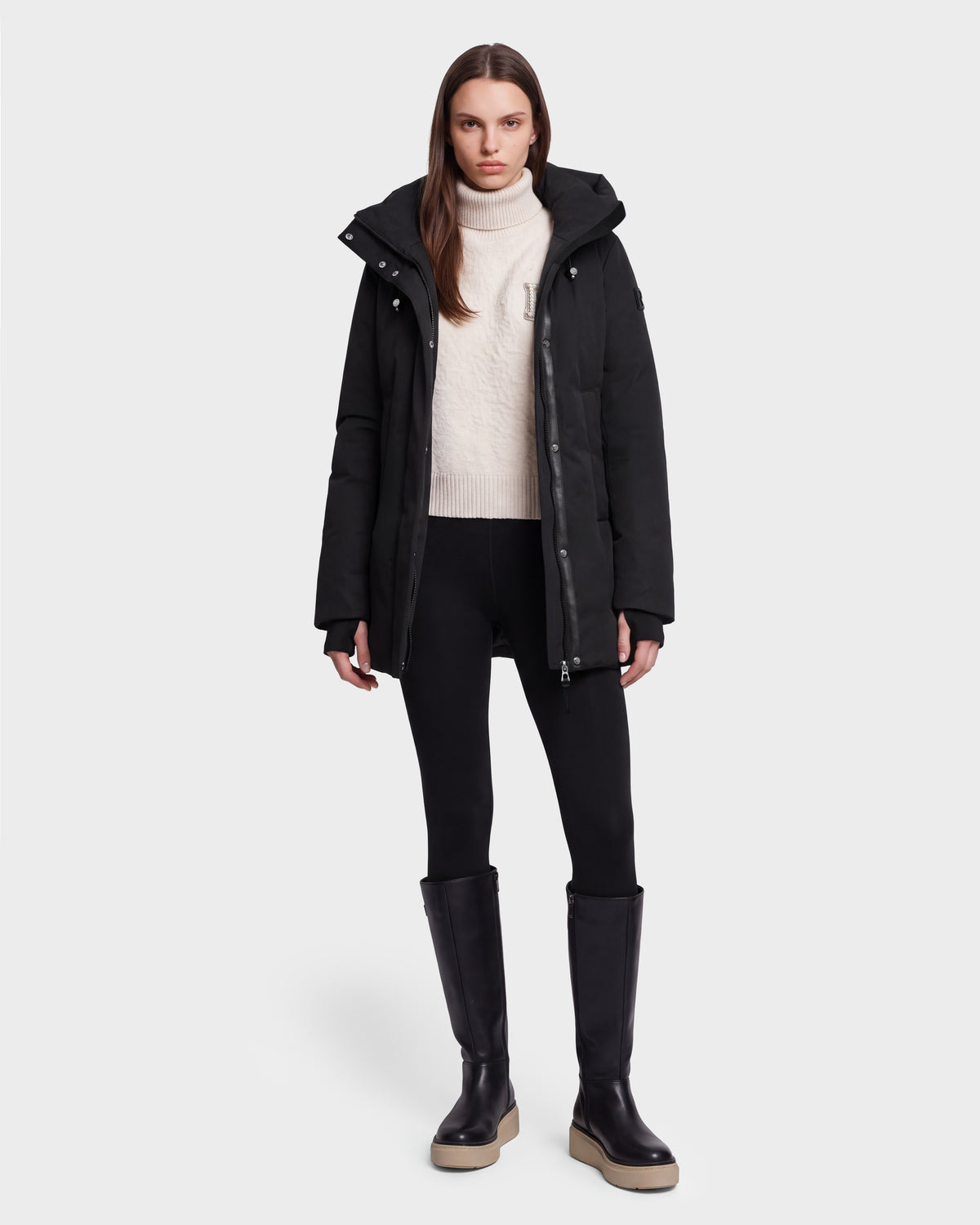 A person wearing the Rudsak OPAL - 8126618 Black winter coat stands facing forward against a plain white background, styled with a white turtleneck sweater, black leggings, and black knee-high boots.
