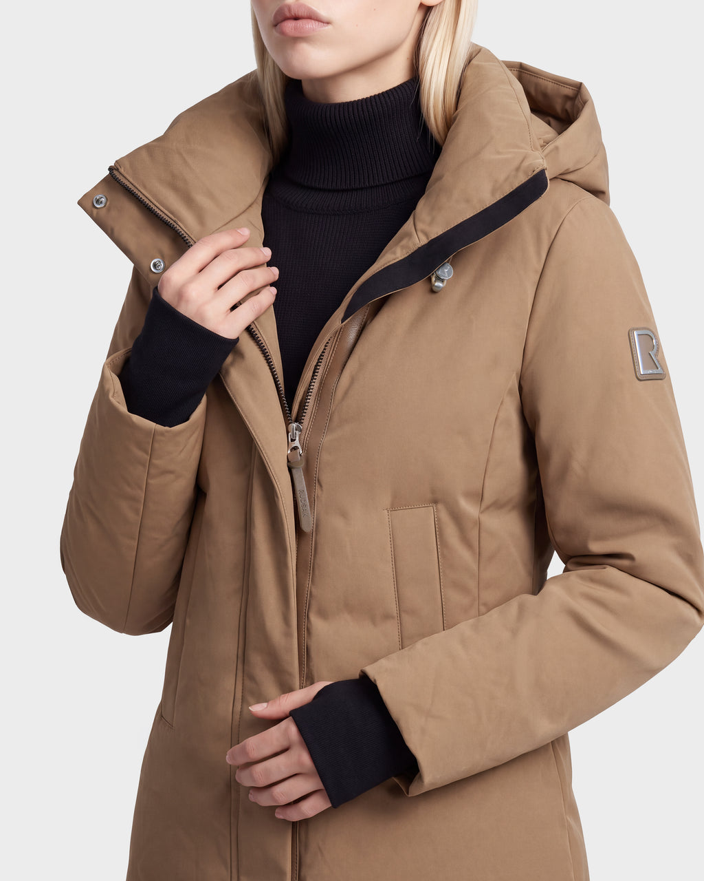 A woman wears the Rudsak OPAL - 8126618 Camel parka over a black turtleneck, one hand on the collar and the other inside her coat. Her face is partially visible.