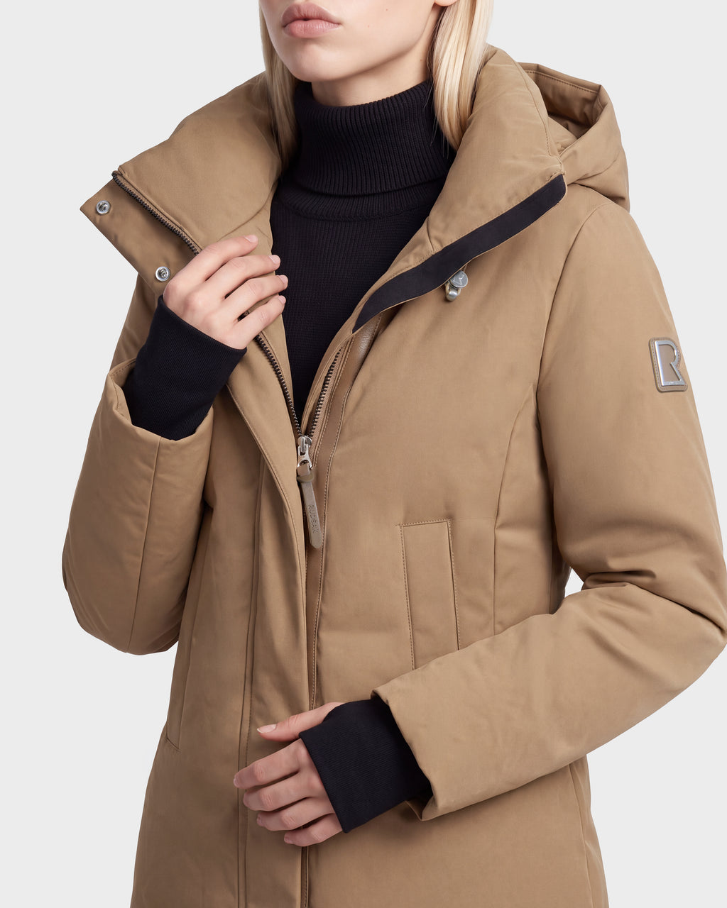 A woman wears the Rudsak OPAL - 8126618 Camel parka, partially unzipped over a black turtleneck, with her hand by the collar and only her lower face and torso in view.