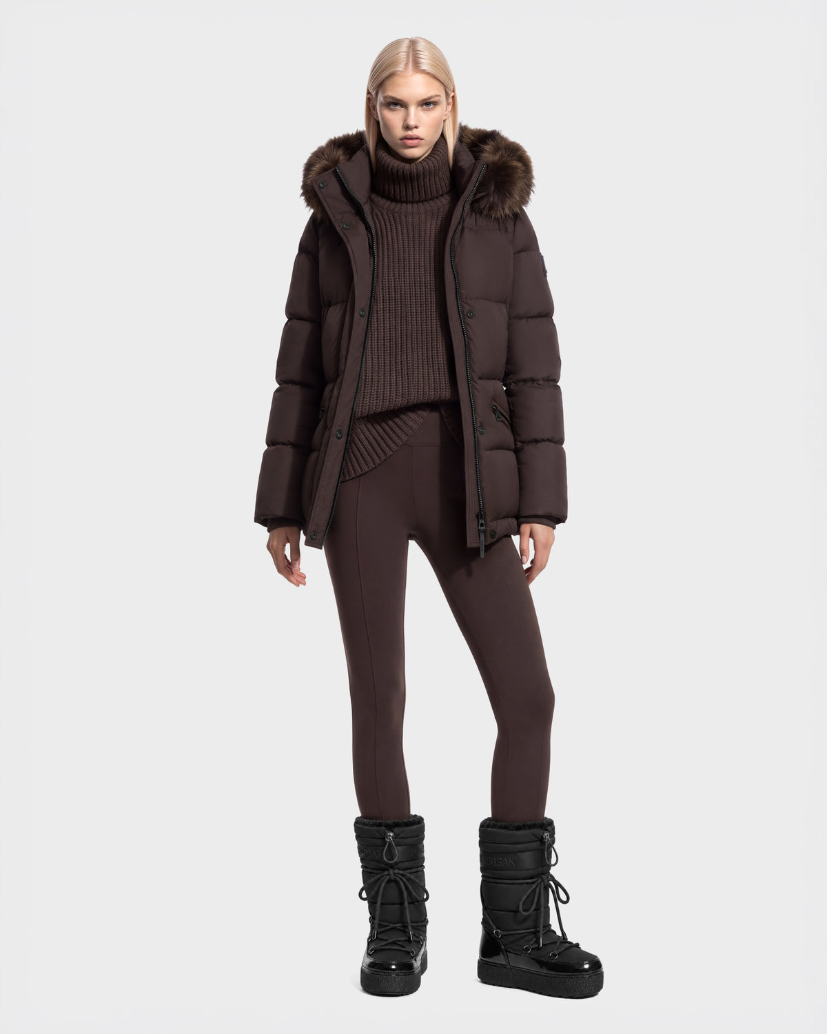 A model wears a Rudsak GRACE FF - 8126535 Espresso dark brown puffer coat with a fur-trimmed hood, a matching chunky turtleneck sweater, dark leggings, and black insulated winter boots.