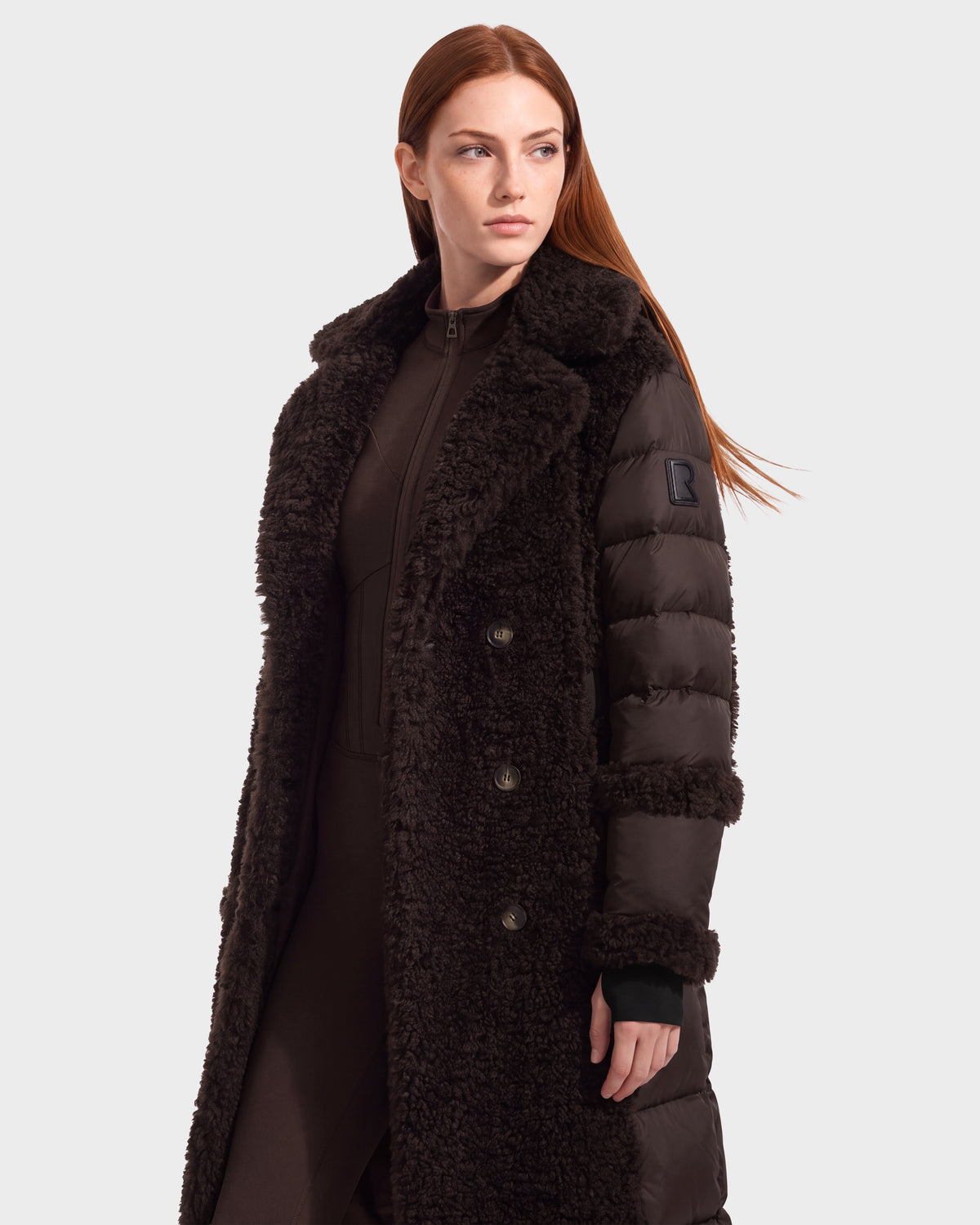 A woman with long red hair wears the Rudsak NOELLE - 8126971 Espresso, a dark double-breasted faux fur coat featuring a textured, fuzzy surface and puffer sleeves. She stands facing left, looking off to the side against a plain background.