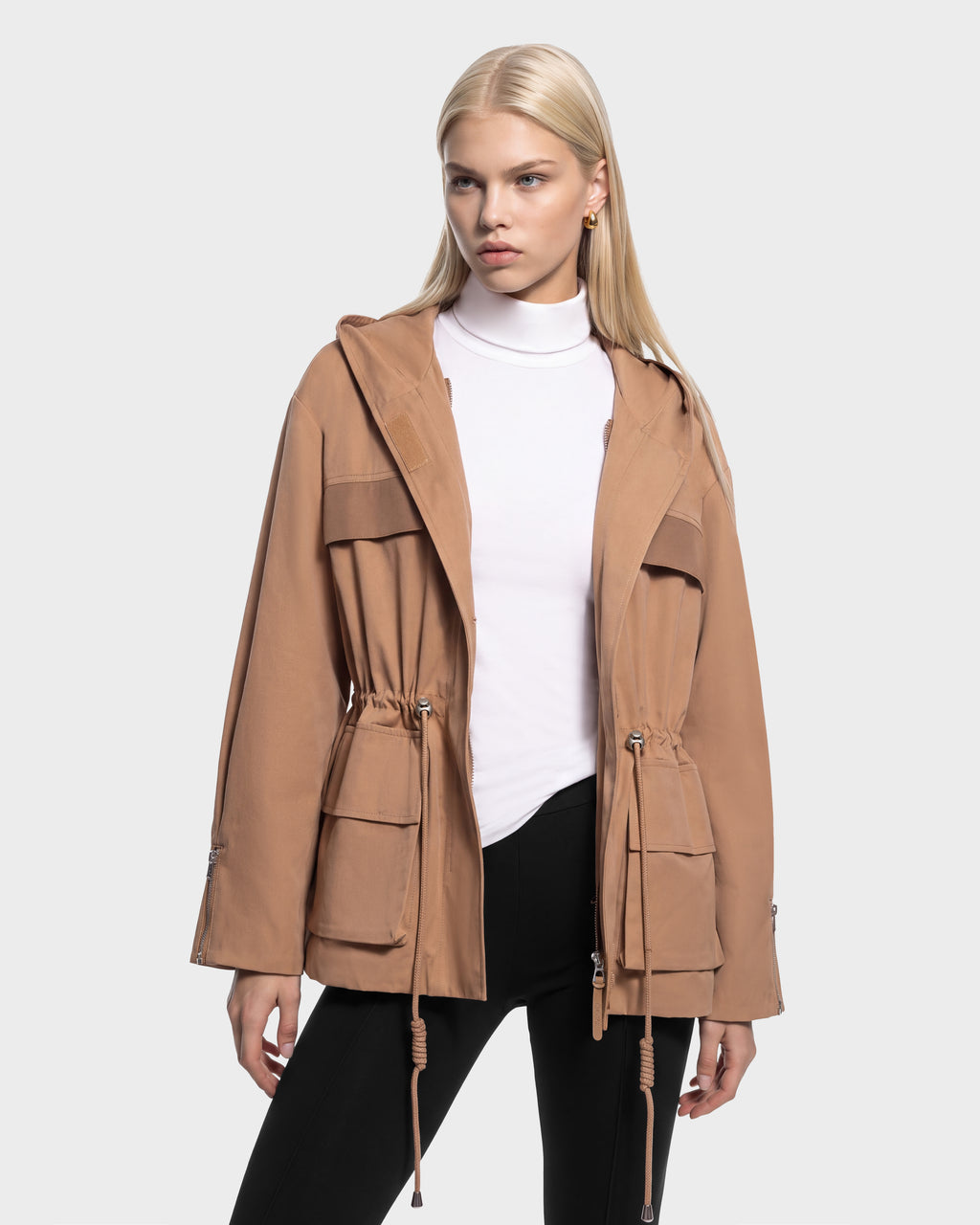 A woman wears a camel-colored RUDSAK ROSALINDA - 8126523 Camel hooded utility jacket over a white turtleneck and black leggings.