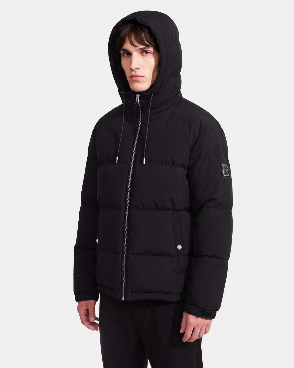 A young man stands against a plain white background, looking straight ahead with a neutral expression while wearing the STERLING EL - 6126759 Black hooded puffer jacket by Rudsak, featuring a front zipper and snap buttons.