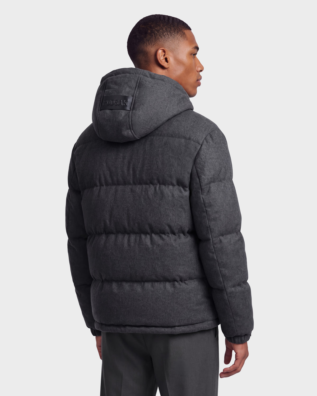 A man stands facing away against a light gray background, showcasing the back of the Rudsak STERLING W - 6126760 Heather Charcoal puffer jacket with a hood.