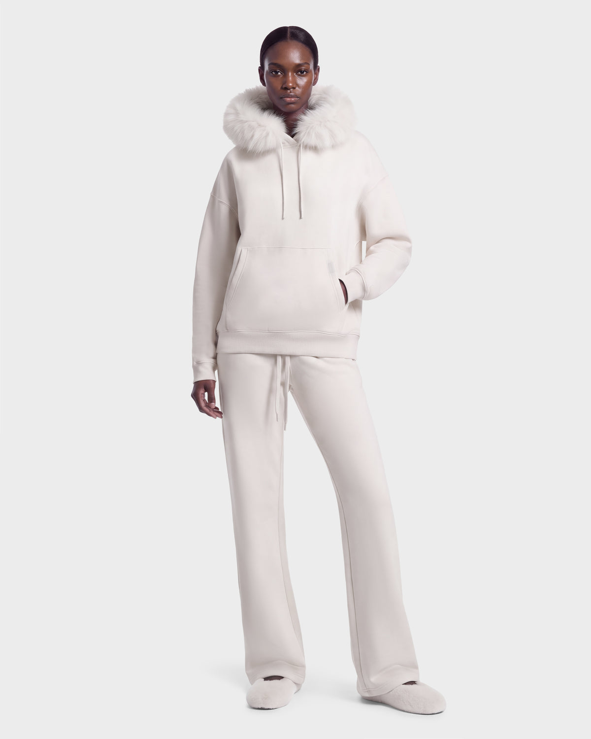 A person wearing the Rudsak ANGEL - 8526725 Vanilla hooded sweatshirt with a fur-trimmed hood, matching stretch cotton sweatpants, and slippers for a refined off-duty look.