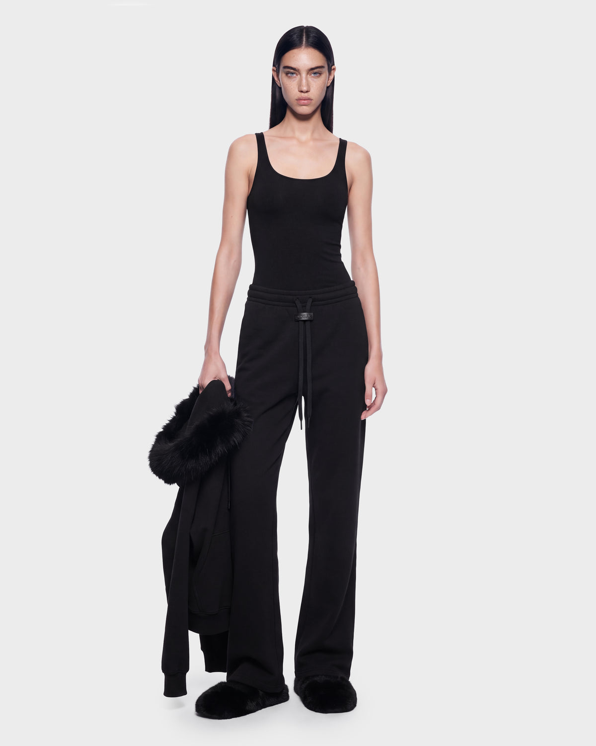 A person wearing a black tank top and contemporary stretch cotton sweatpants holds a black ANGEL - 8526725 Black jacket with a fur-trimmed hood while wearing black slippers.