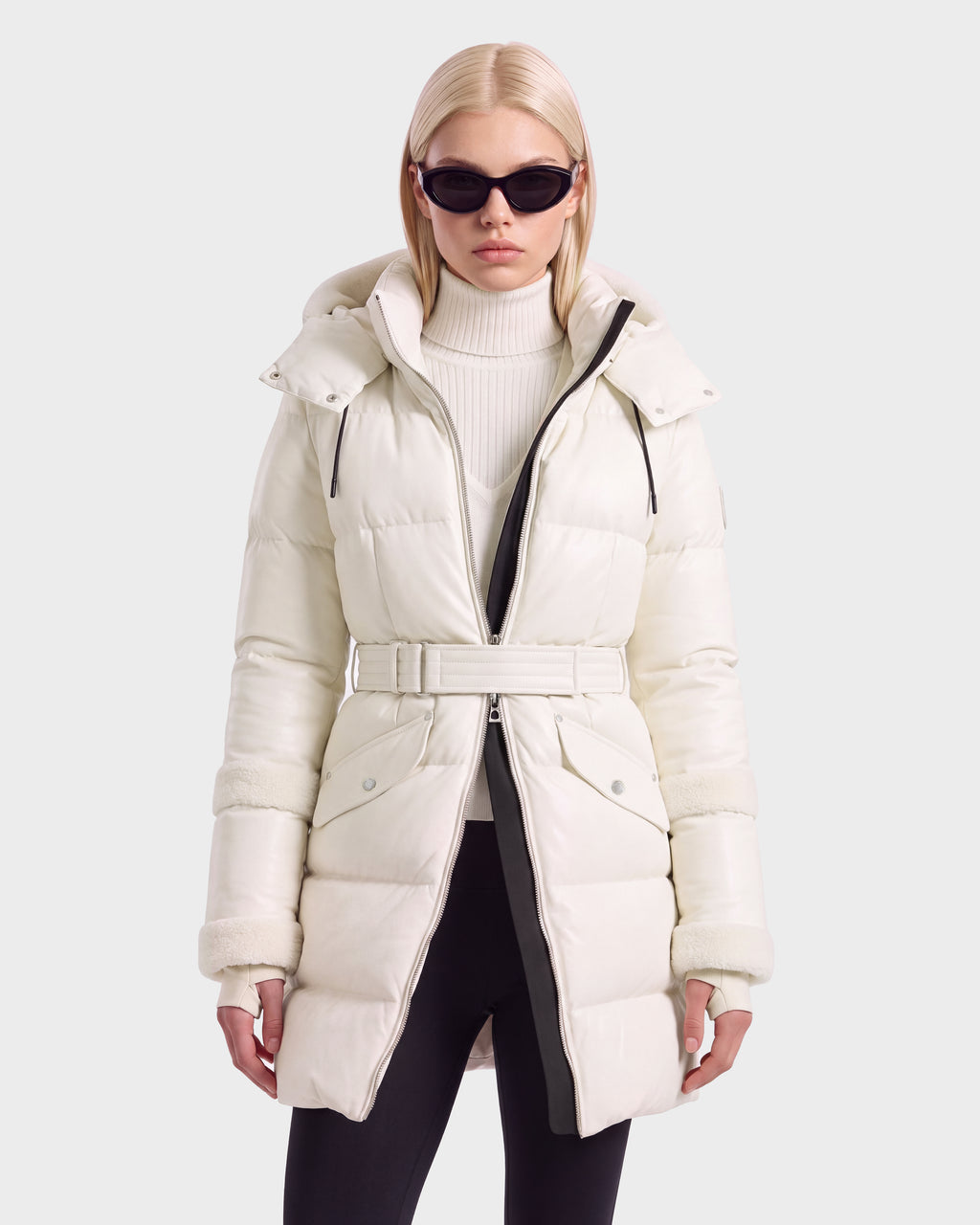 A person wearing a vanilla-colored Rudsak AUDREY L - 8124585 belted down puffer jacket with a hood and black sunglasses.