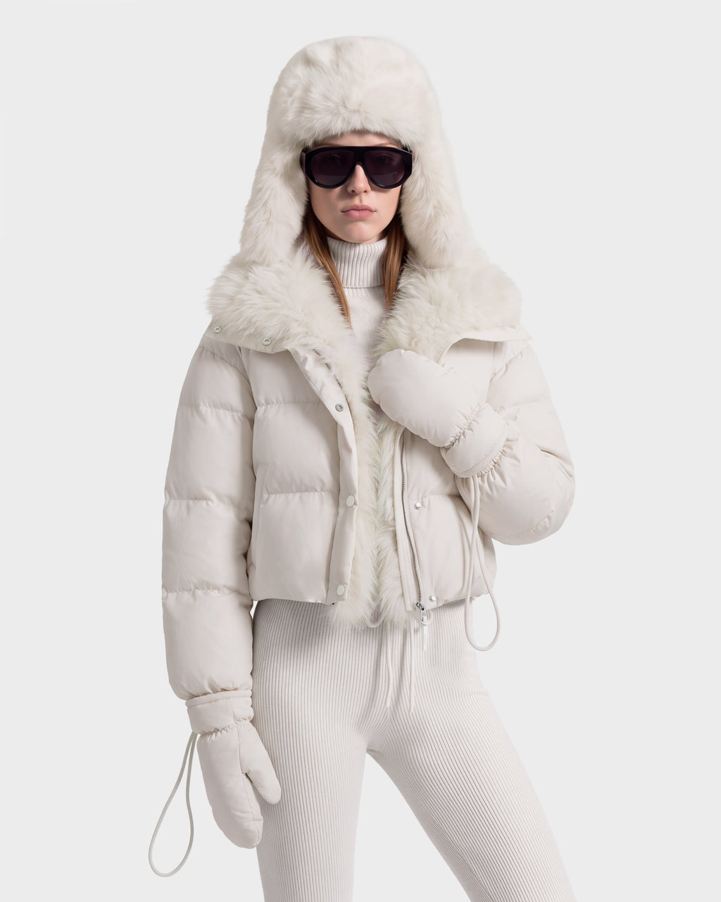A person wearing a Rudsak ALONDRA - 8126992 Vanilla RDS-certified down cropped puffer jacket with a faux-fur trimmed hood, matching white turtleneck, ribbed leggings, and mittens, accessorized with dark sunglasses.