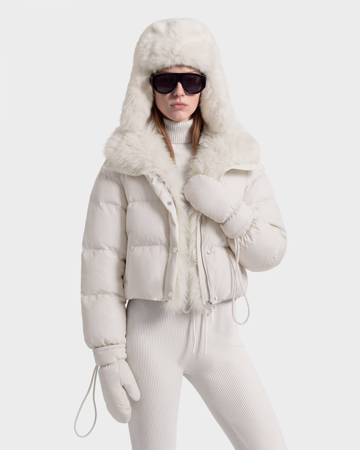 A person wearing a Rudsak ALONDRA - 8126992 Vanilla RDS-certified down cropped puffer jacket with a faux-fur trimmed hood, matching white turtleneck, ribbed leggings, and mittens, accessorized with dark sunglasses.