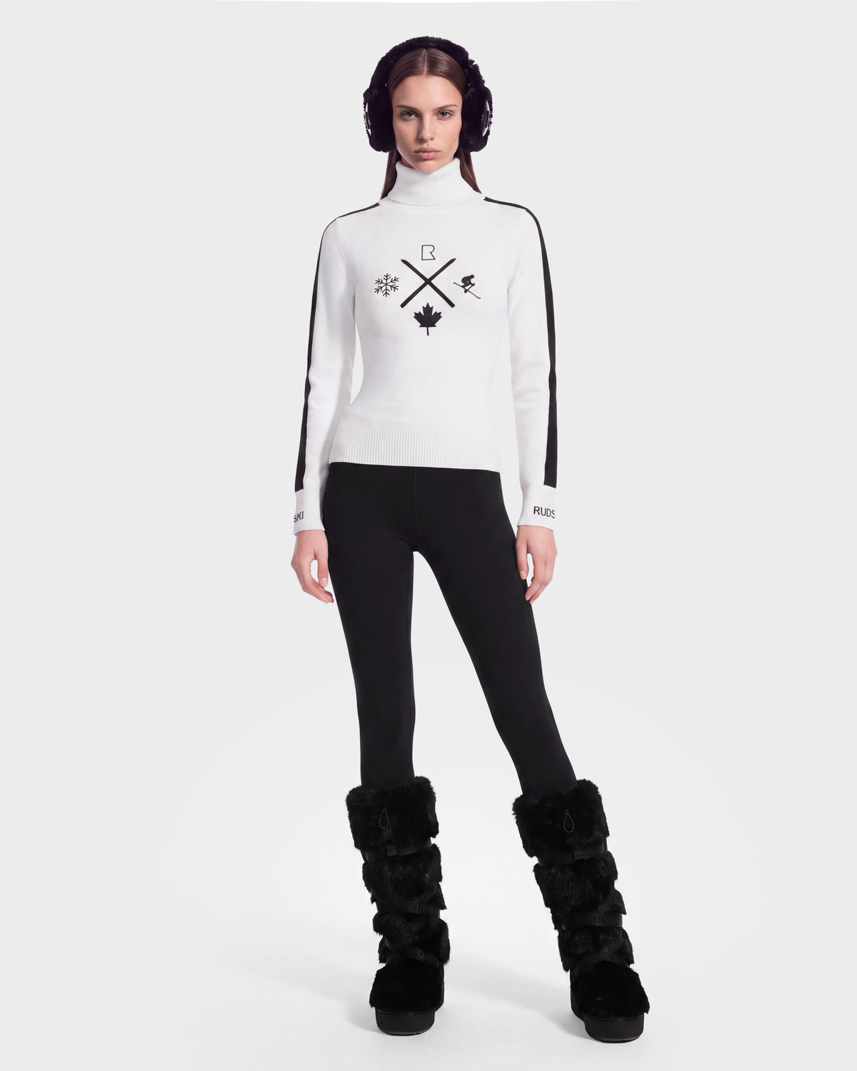 A woman stands facing forward against a plain white background, wearing the Rudsak ELIZABETH - 8524623 Optic White women’s ski turtleneck sweater with black graphics, paired with black leggings, black furry boots, and black earmuffs—an effortlessly chic choice for any winter wardrobe or après-ski outing.