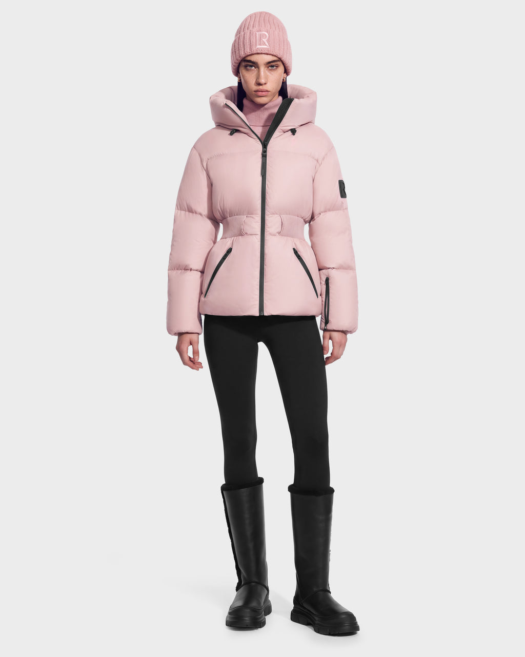 A person wearing a Rudsak DIAMOND - 8126772 Vintage Rose pale pink quilted puffer jacket with a belted waist, matching beanie, black leggings, and black knee-high boots.