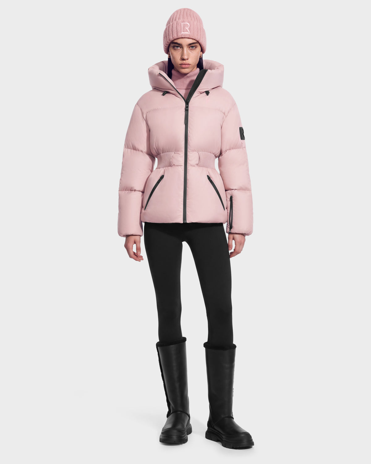 A person wearing a Rudsak DIAMOND - 8126772 Vintage Rose pale pink quilted puffer jacket with a belted waist, matching beanie, black leggings, and black knee-high boots.