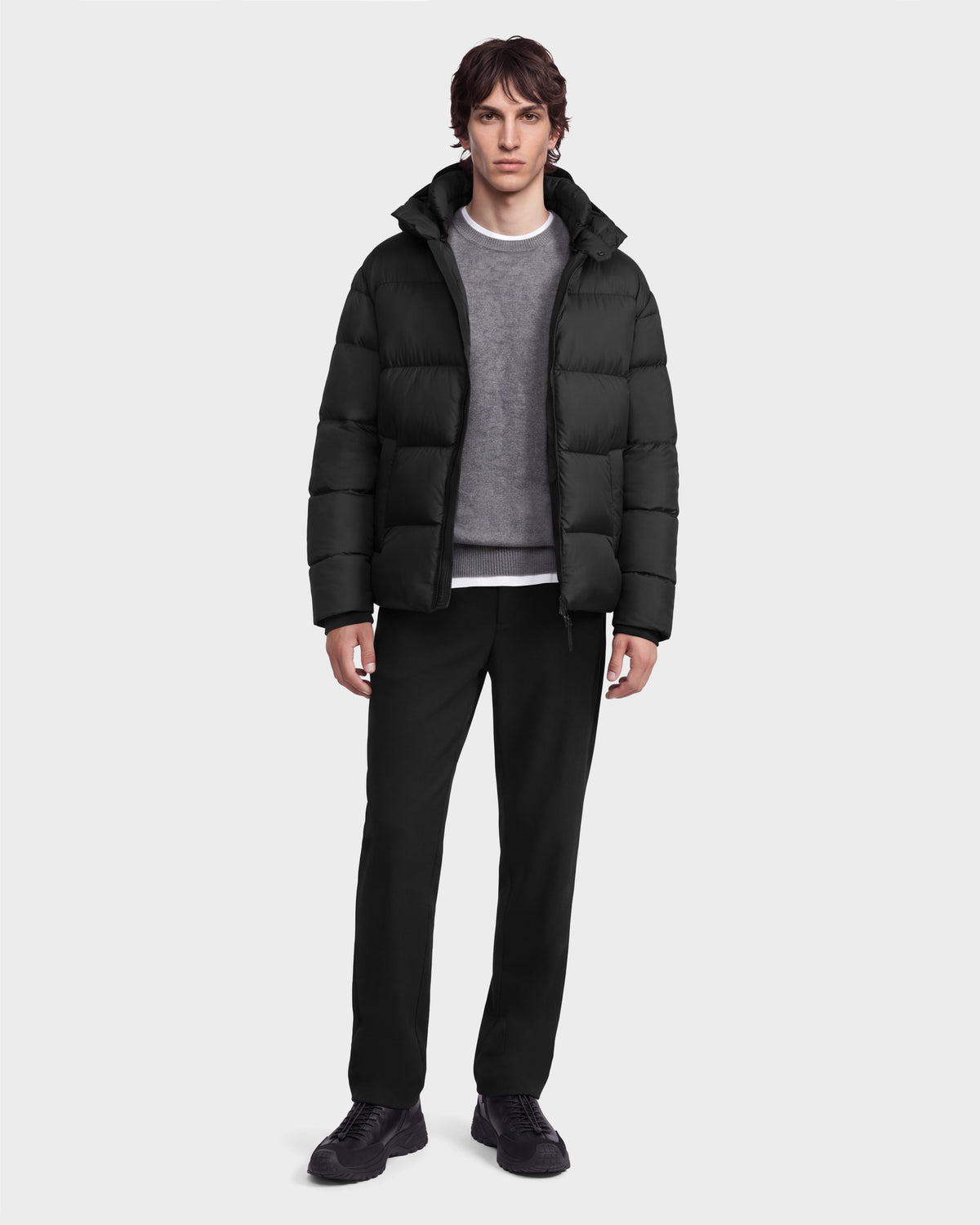 A man wearing a black Rudsak JASON - 6126803 Black hooded winter puffer jacket over a grey sweater and black trousers.