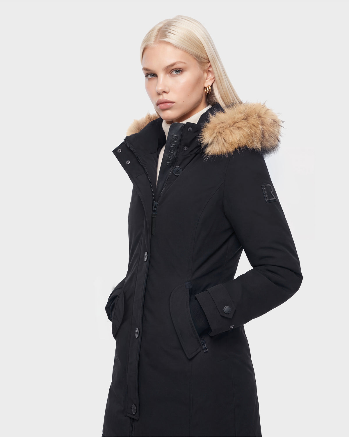 A woman with straight blonde hair models the KEYSHIA - 8126517 Black by Rudsak, a black down parka with a fur-trimmed hood, hands in pockets and facing slightly to the side against a white background.