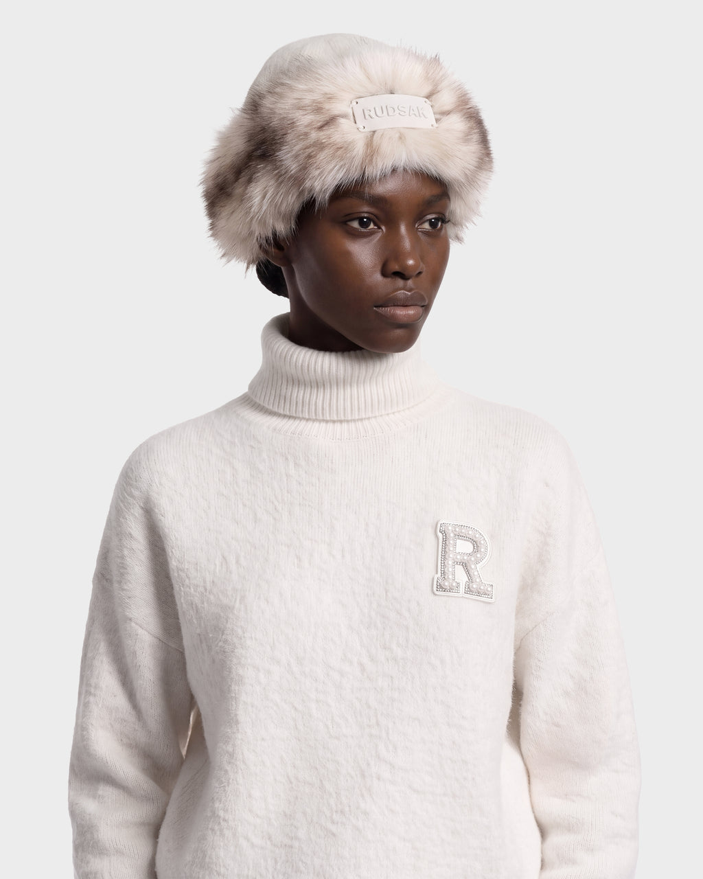 A person wears a white LORETTA W - 8426510 Vanilla turtleneck sweater with a rhinestone R patch and a RUDSAK faux-fur bucket hat with a centered logo plaque.