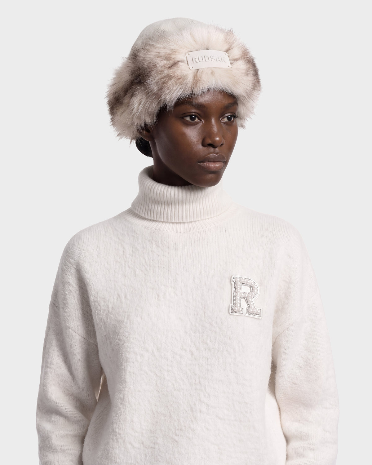 A person wears a white LORETTA W - 8426510 Vanilla turtleneck sweater with a rhinestone R patch and a RUDSAK faux-fur bucket hat with a centered logo plaque.