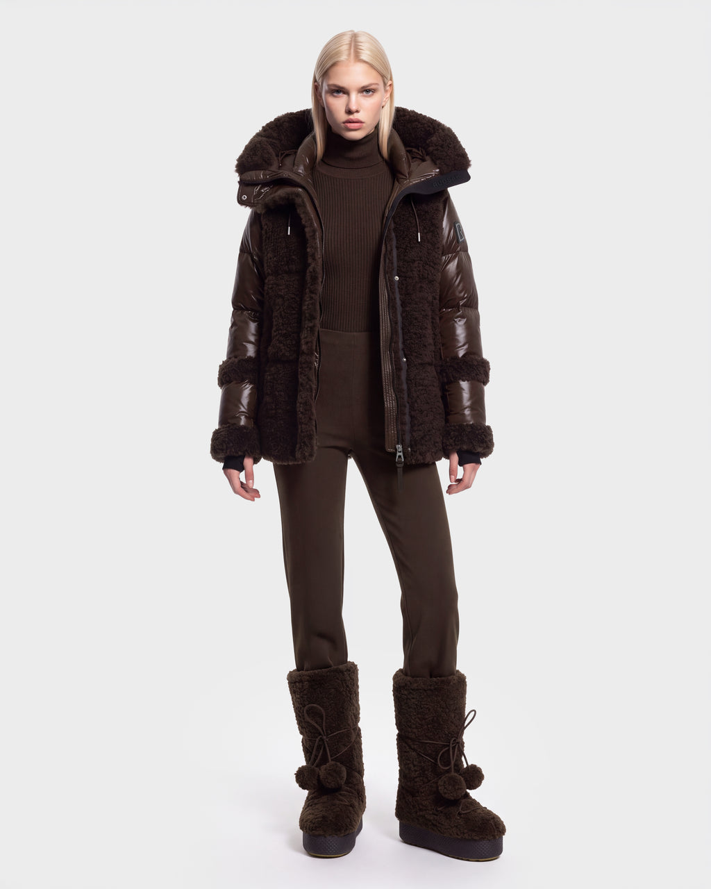 A person stands facing forward, wearing the Rudsak MARISSA - 8126728 Espresso dark brown down insulated puffer coat with fur trim, paired with matching brown pants and large brown fur boots—perfect winter outerwear. The background is plain white.