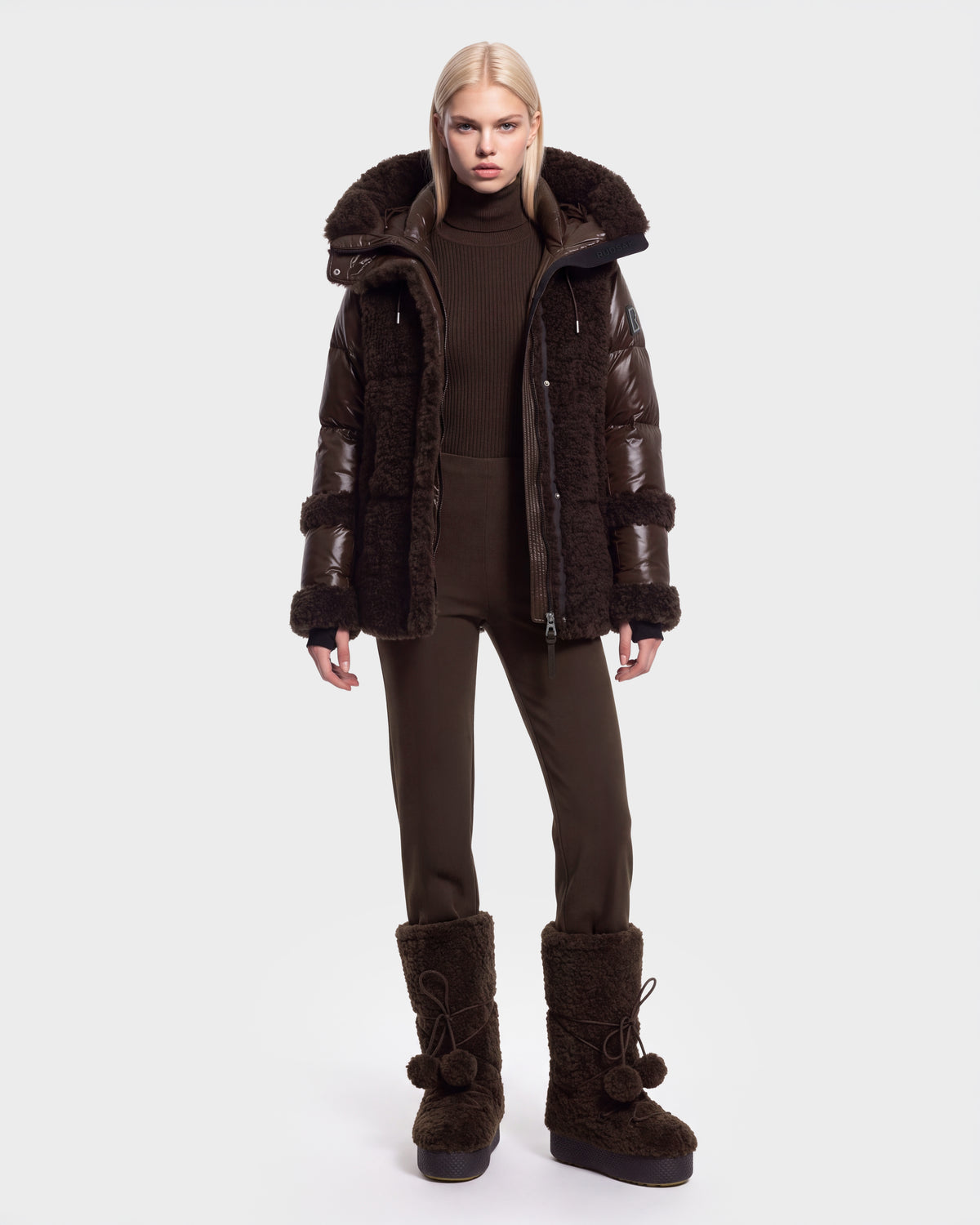 A person stands facing forward, wearing the Rudsak MARISSA - 8126728 Espresso dark brown down insulated puffer coat with fur trim, paired with matching brown pants and large brown fur boots—perfect winter outerwear. The background is plain white.