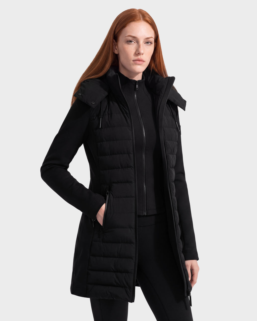 A person with long red hair wears the Rudsak MERINA - 8125056 Black quilted down jacket, which is knee-length with a zipper and pockets, standing against a plain light gray background.