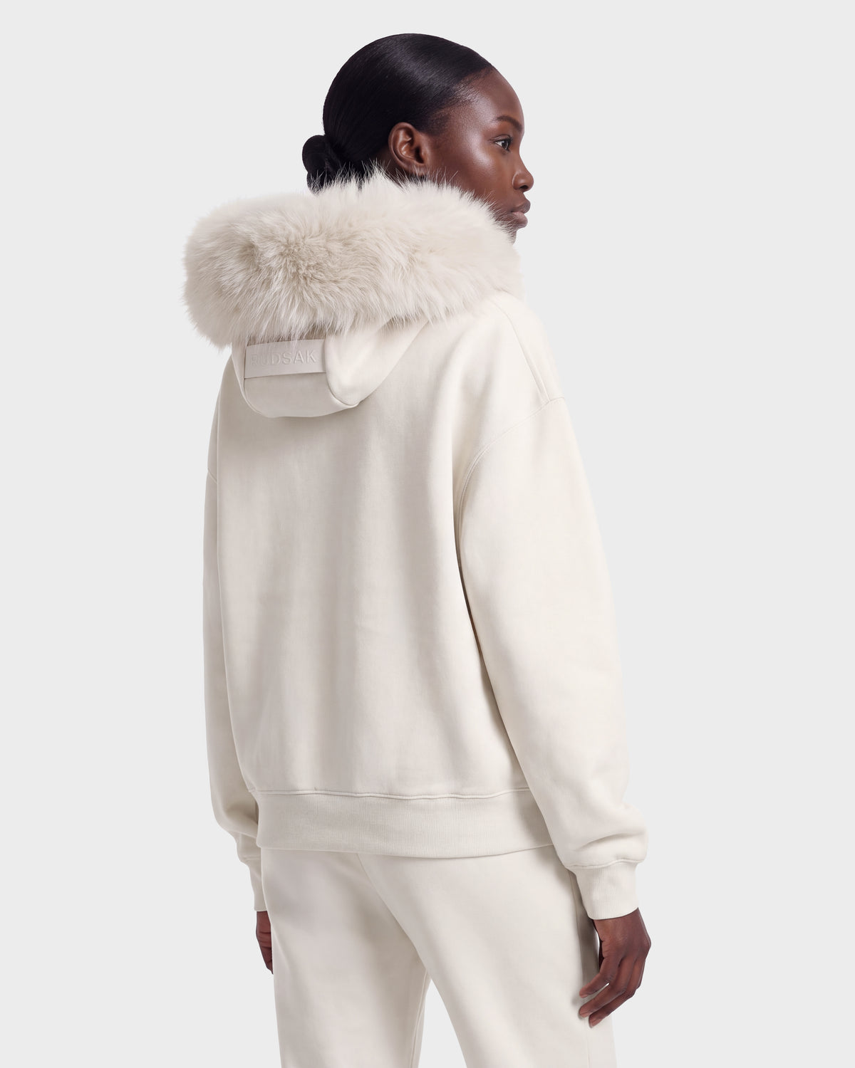 A person wearing the Rudsak MILENA - 8526689 Vanilla, an off-white hoodie with a fur-trimmed hood and matching pants, stands against a plain light background, facing away.