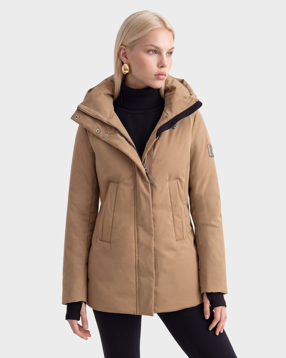 A woman with blonde hair wears the Rudsak OPAL - 8126618 Camel parka over a black turtleneck and pants, standing against a plain light background.