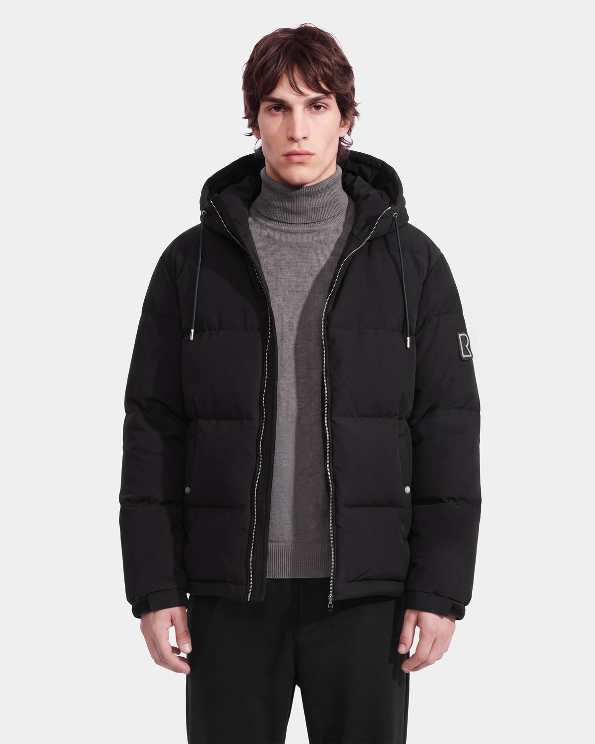 A person stands facing forward, wearing the Rudsak STERLING EL - 6126759 Black puffer jacket with a hood over a gray turtleneck sweater and black pants, against a plain light background.