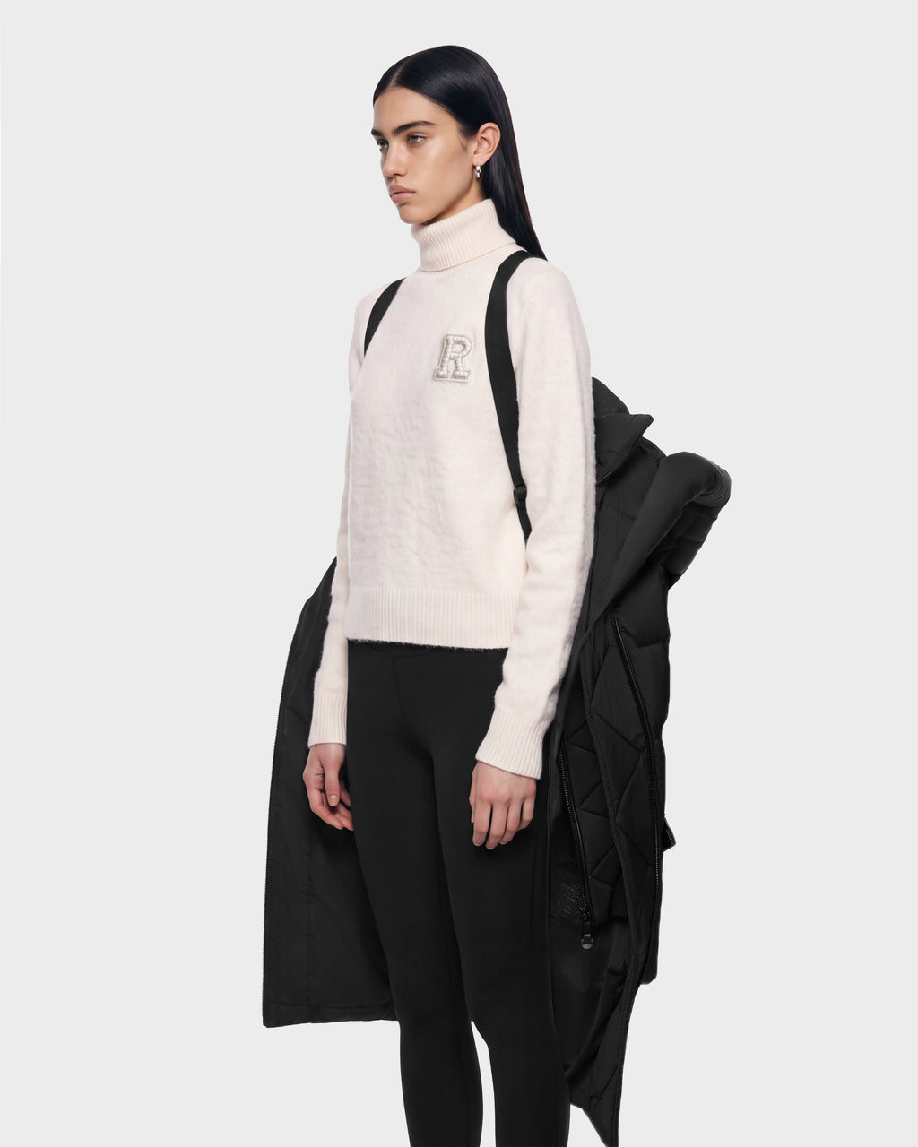 A person wears a cream-colored turtleneck sweater with a prominent letter R emblem and black leggings, layered with a TAYLOR M - 8125965 Black jacket draped over their shoulders.