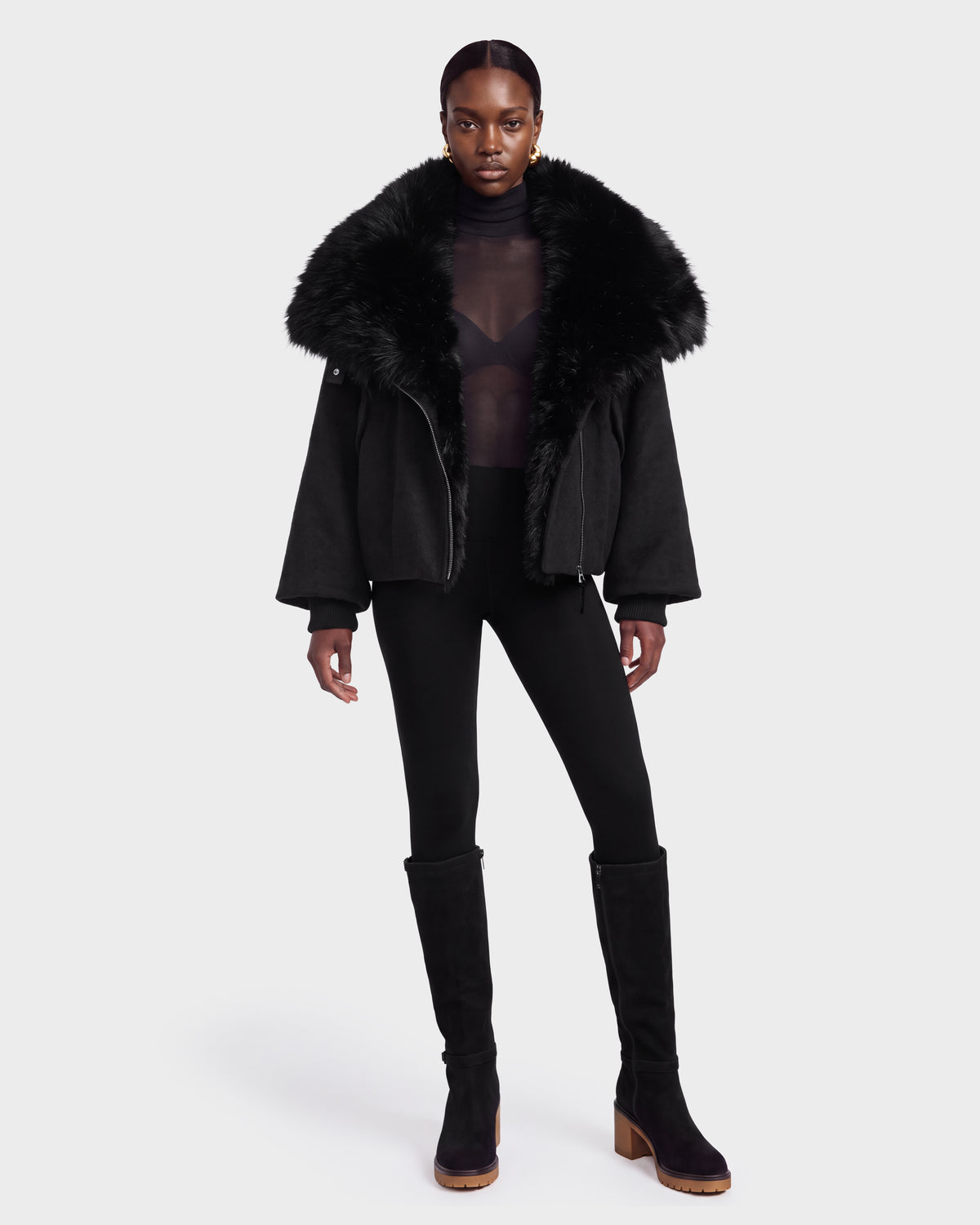 A person wearing the Rudsak ZARIA - 8126999 Black jacket, featuring a large faux fur collar, styled with a sheer black top, black leggings, and black knee-high boots stands against a plain white background.