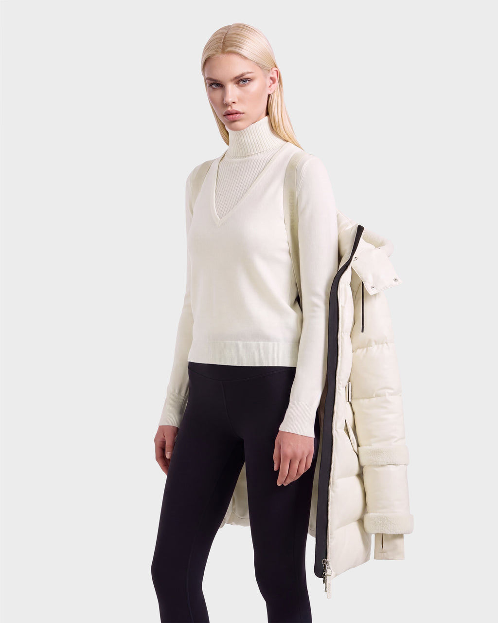 A woman wearing a cream turtleneck sweater and black leggings, with a cream Rudsak AUDREY L - 8124585 Vanilla down puffer jacket draped over her shoulders.